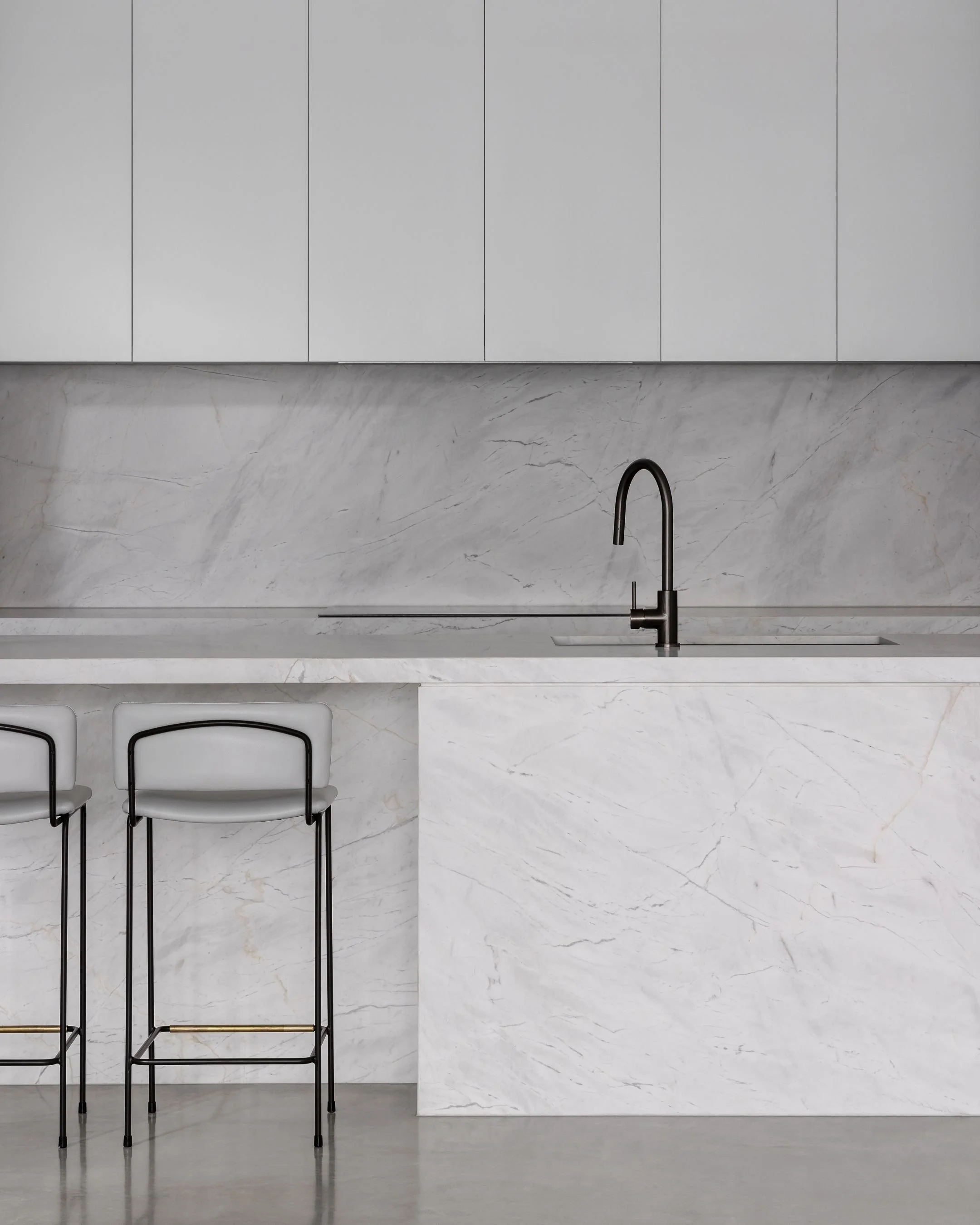 Minimalist kitchen with white marble countertops, black faucet, closed wall cabinets, and two black and white bar stools.