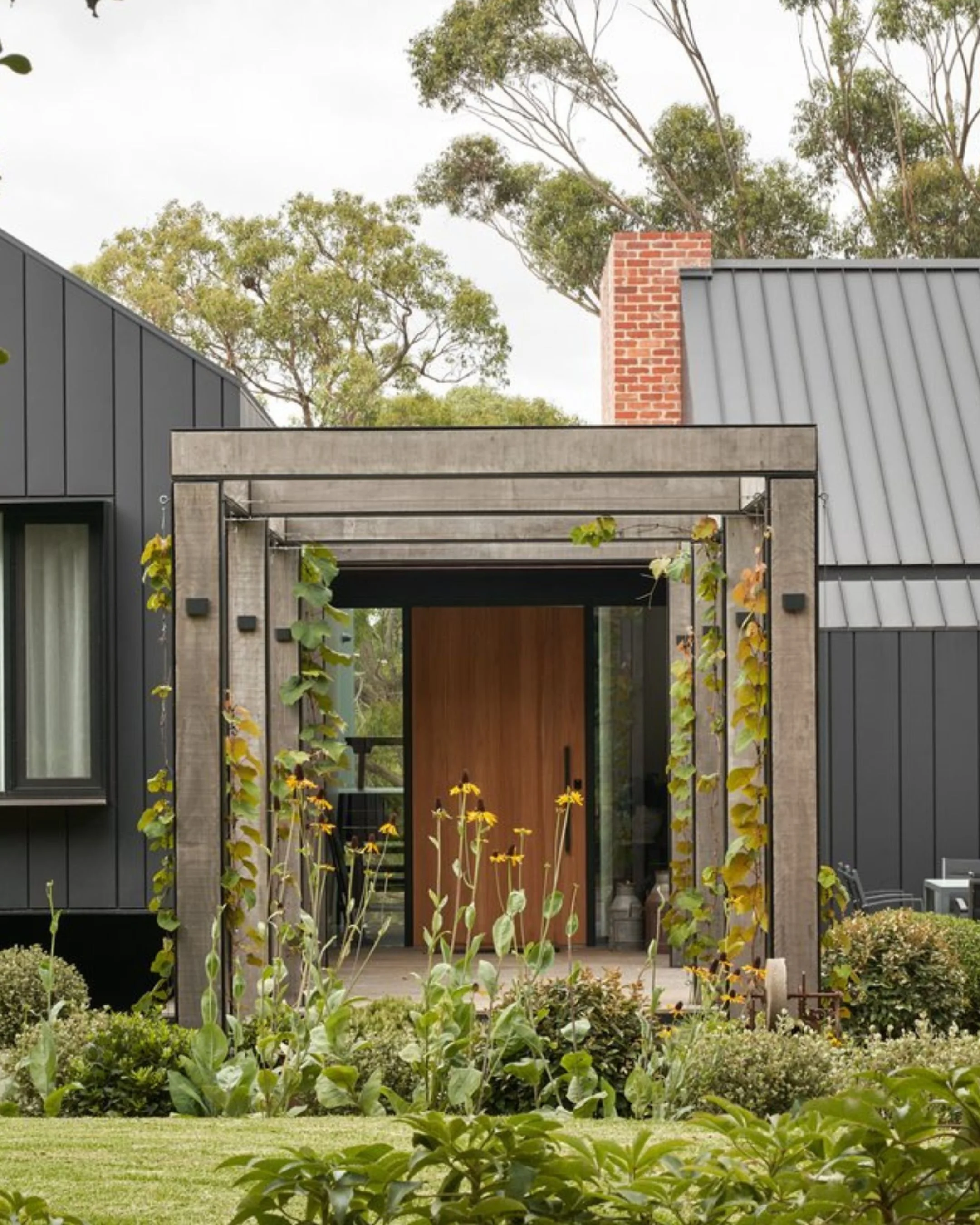 Modern house entrance with wooden door, surrounded by greenery, plants, and outdoor seating.
