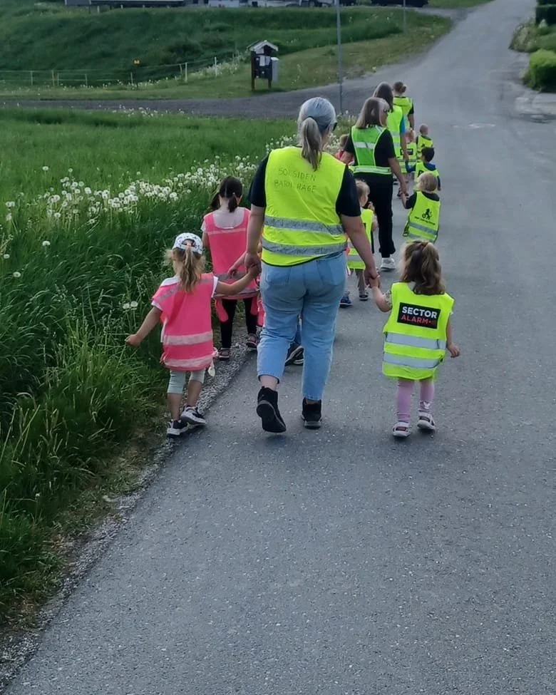 Barn og voksne iført refleksvester går tur langs en vei i landlige omgivelser.
