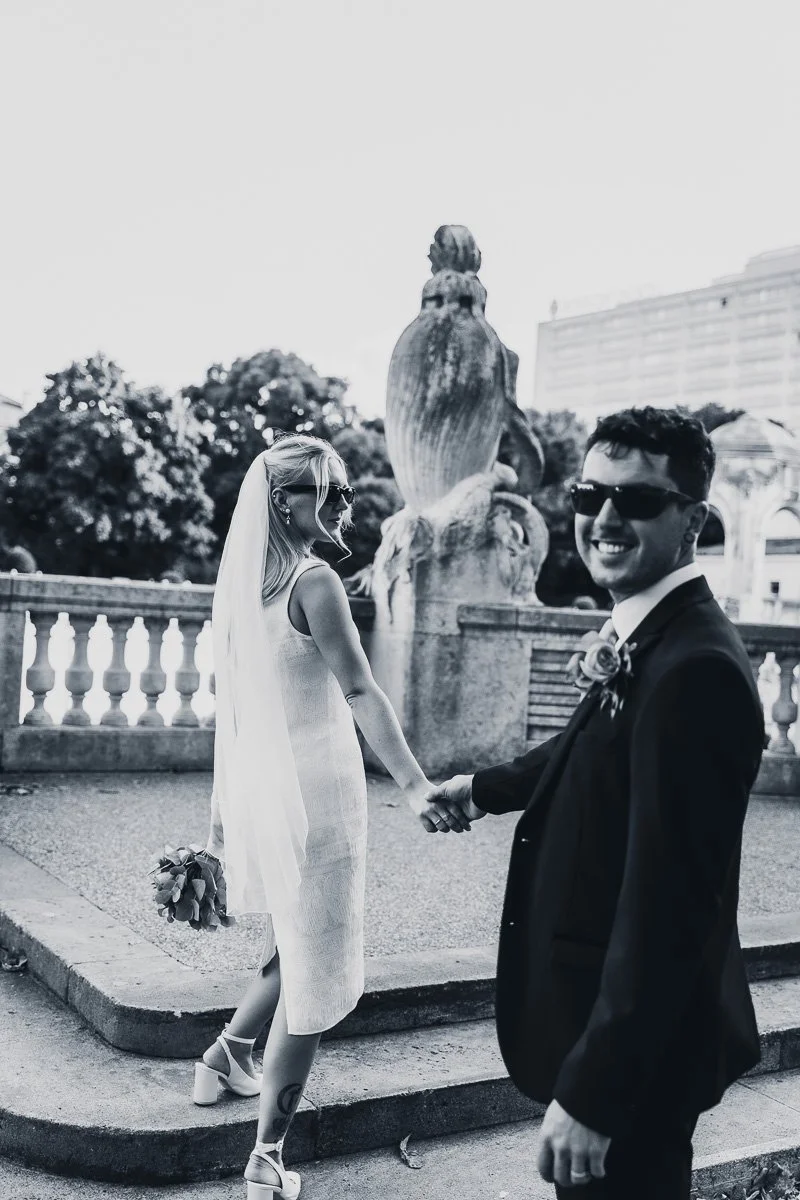 Ein Mann und eine Frau, die sich an einem öffentlichen Ort an einer Brüstung halten, mit einem Brunnen im Hintergrund, in Schwarz-Weiß-Fotografie, anscheinend bei einer besonderen Gelegenheit, alle tragen dunkle Sonnenbrillen.