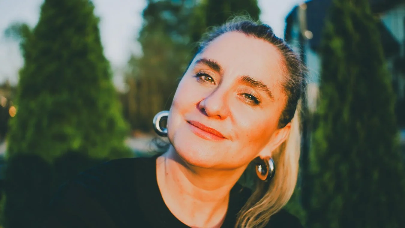 Nahaufnahme eines lächelnden Frauengesichts mit Lightbox in einer grünen, natürlichen Umgebung bei Sonnenuntergang, trägt große Ohrringe und einen schwarzen Pullover.