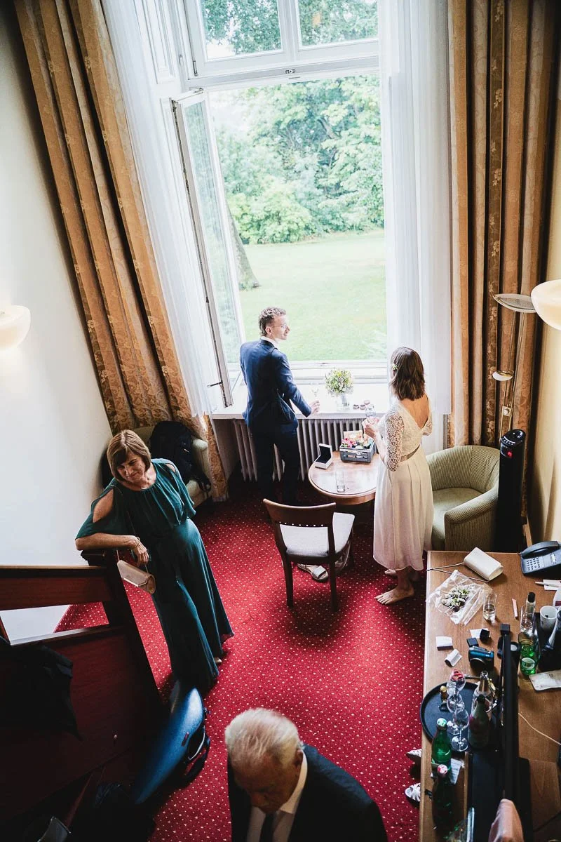 Ein Hochzeitspaar steht vor einem großen offenen Fenster mit einem Blick auf einen grünen Garten, während eine Frau im dunkelgrünen Kleid und ein älterer Mann im Anzug im Raum sind.