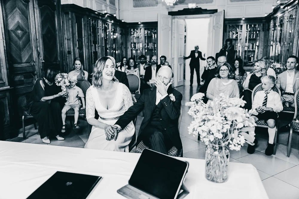 Hochzeitsszene mit Braut und Bräutigam in einem eleganten Raum, umgeben von Gästen, einige lachen und unterhalten sich, im Vordergrund sind Blumen und elektronische Geräte auf einem Tisch.
