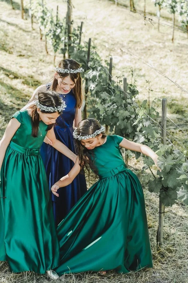 Vier junge Mädchen in Abendkleidern und Blumenkränzen besichtigen eine Weinrebe in einem Weingarten im Freien.