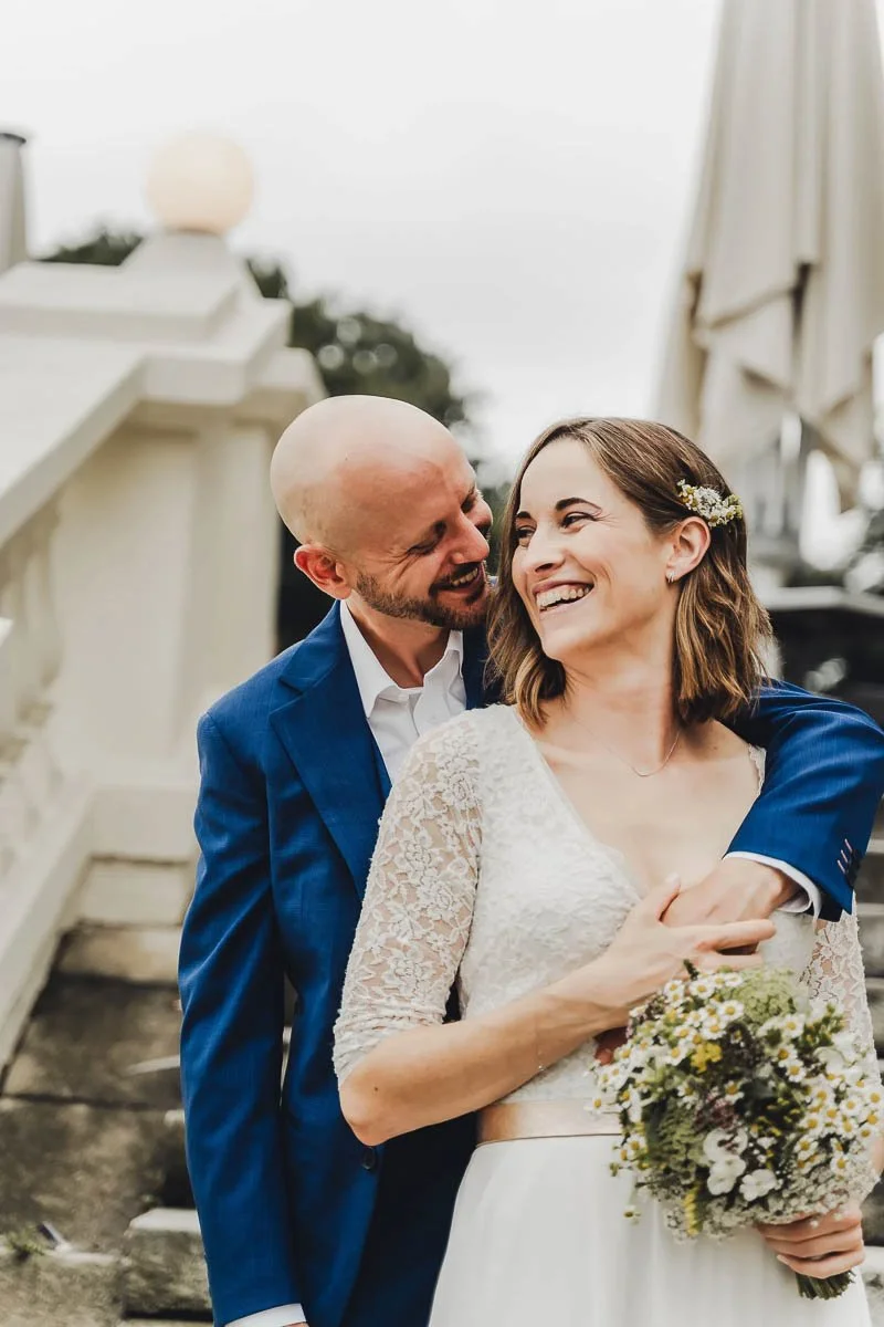 Ein glückliches Brautpaar bei einer Hochzeit im Freien, die Braut trägt ein weißes Kleid mit Spitzenärmeln und hält einen Blumenstrauß, der Bräutigam trägt einen blauen Anzug.