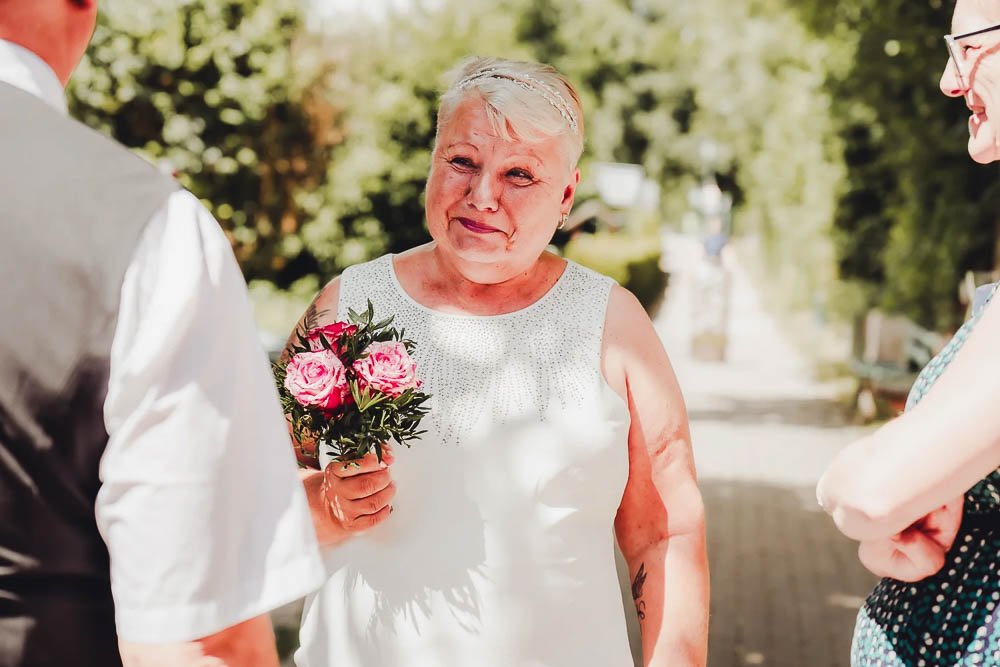 Ältere Frau mit Kurzhaarfrisur, in weißem Kleid, hält einen Blumenstrauß aus pinken Rosen, spricht mit zwei anderen Personen im Freien an einem sonnigen Tag.