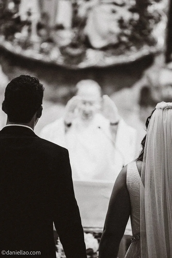 Ein Brautpaar bei einer Hochzeit, mit dem Priester im Hintergrund, in Schwarz-Weiß aufgenommen.