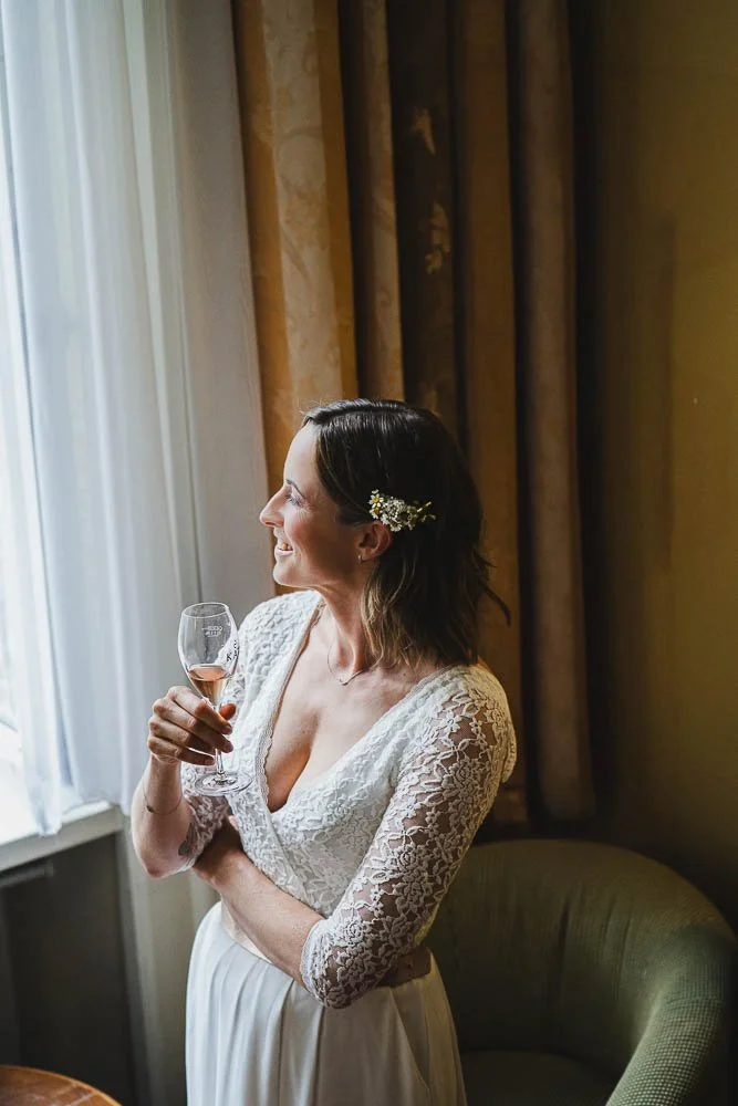 Eine Frau in einem weißen Spitzenkleid hält ein Weinglas und steht neben einem Fenster mit Vorhängen, die im Licht leuchten.