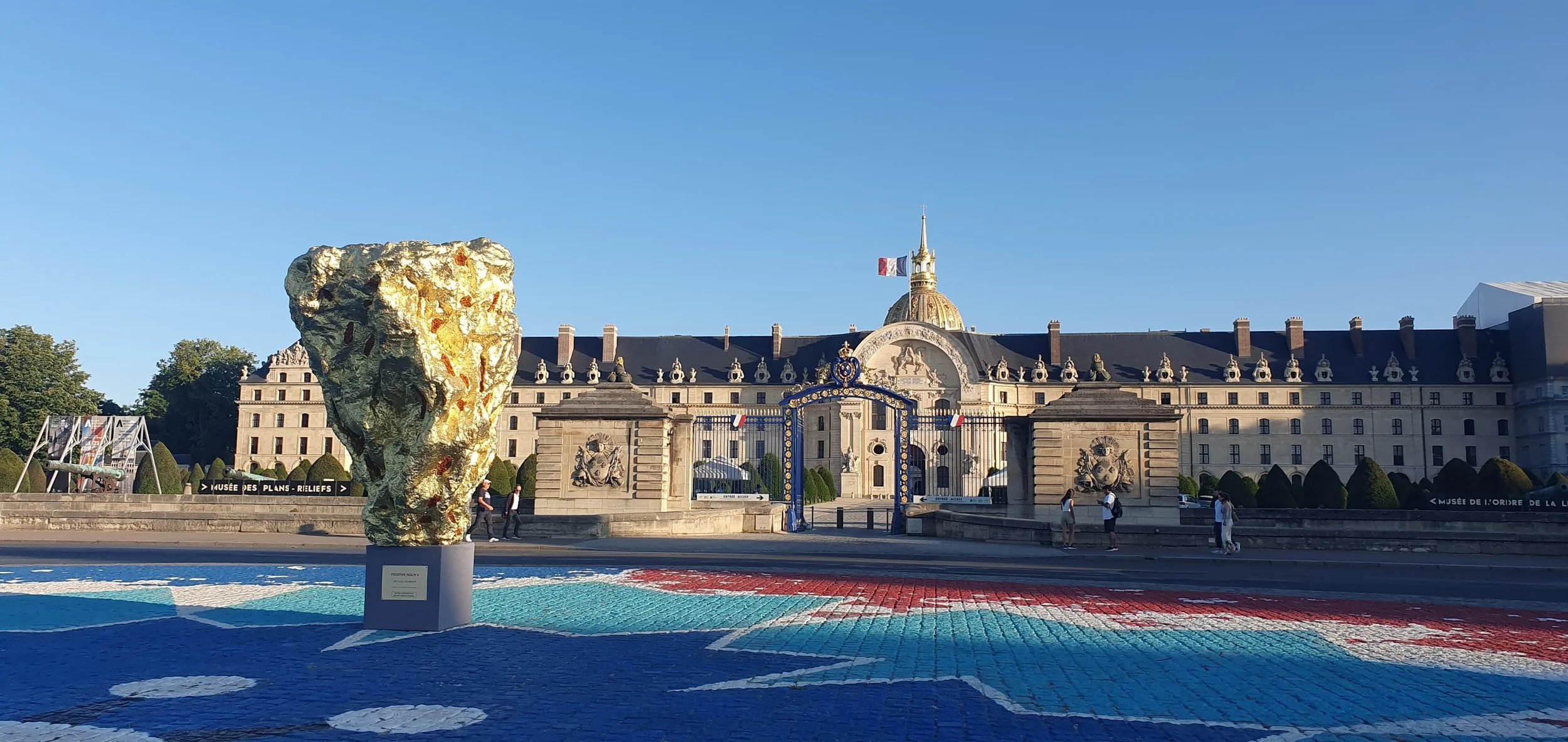 Large abstract bronze sculpture made of textured gold, baltic amber and solid gold, located Place des invalides in Paris. People are walking nearby on a sunny day.