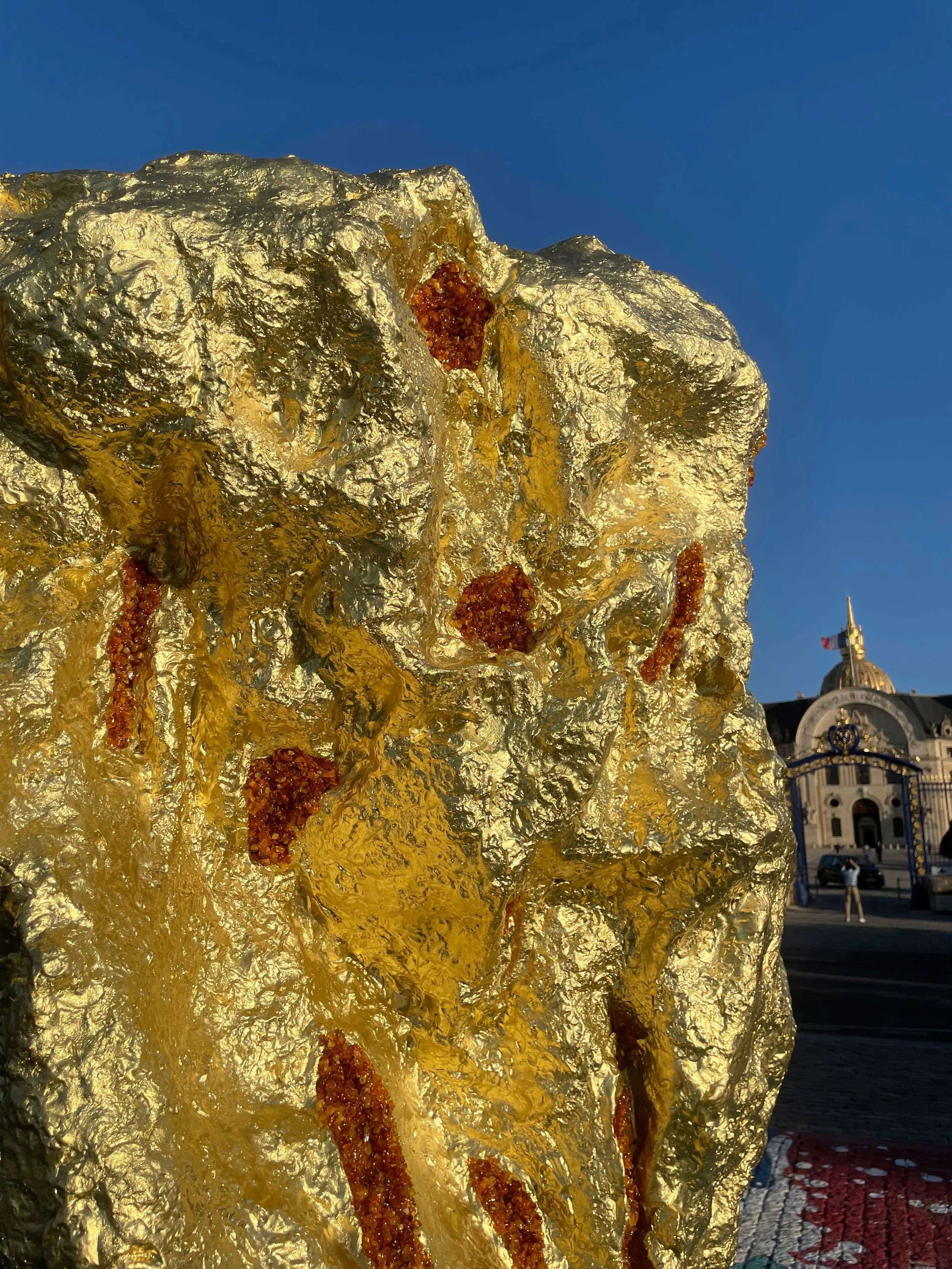 Large abstract bronze sculpture made of textured gold, baltic amber and solid gold, located Place des invalides in Paris, on a sunny day.