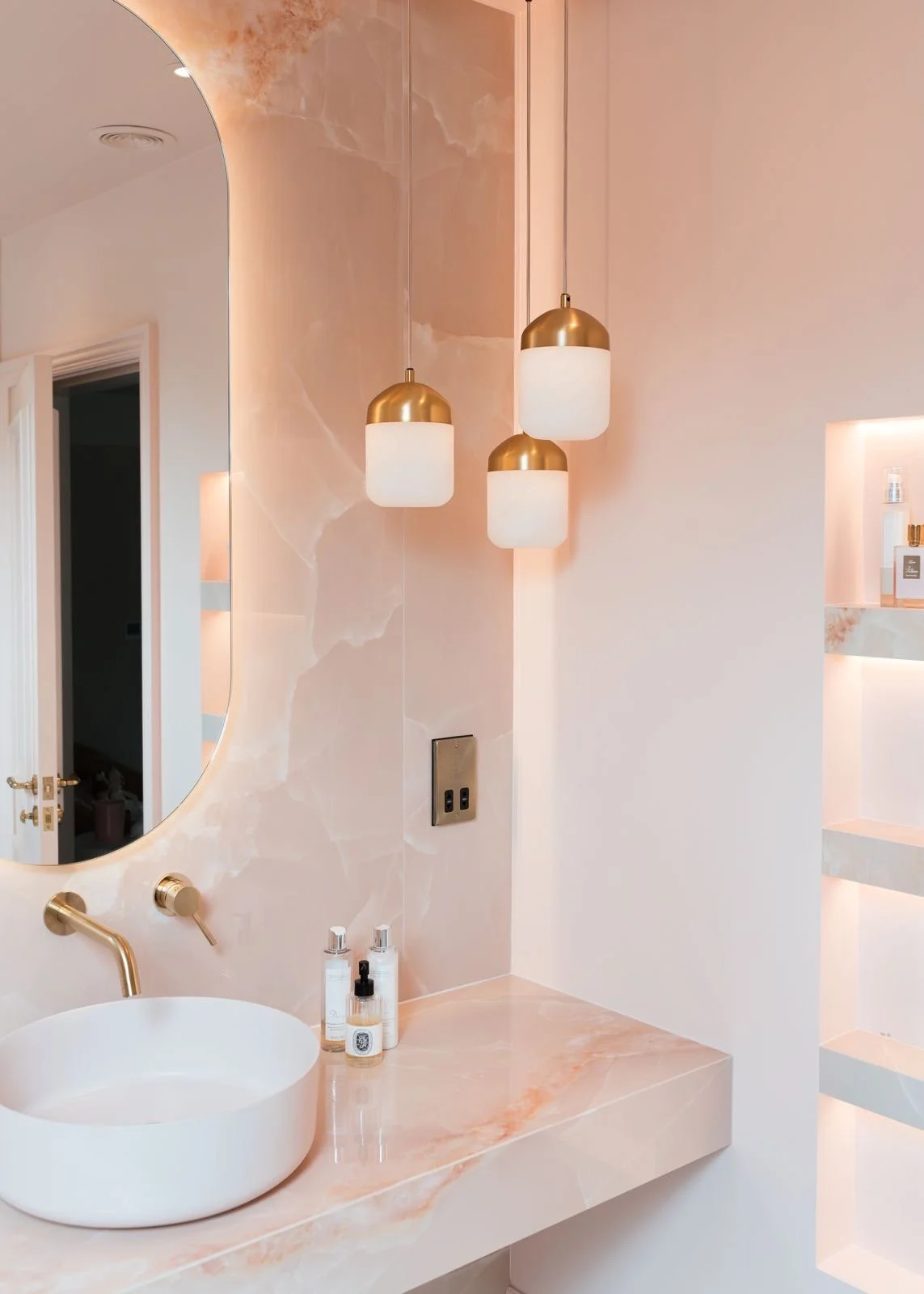 Pink onyx large format tile.  Luxury bespoke vanity. Counter top basin. Wall mounted brass taps and faucet. Recessed shelves with mitred edges. Luxury bathroom.