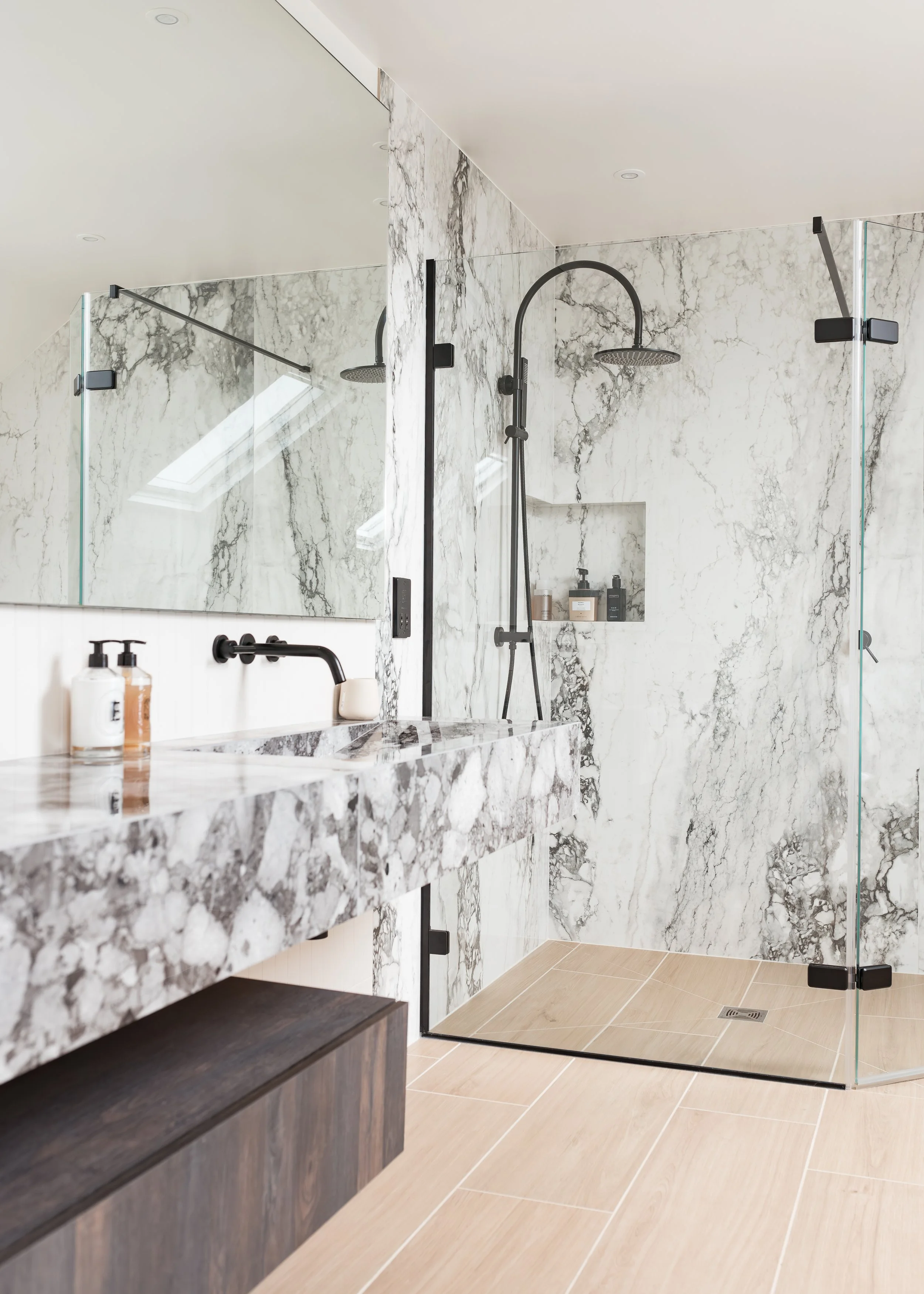 Modern bathroom with marble walls, a glass shower enclosure, a marble countertop with black faucet, and beige tiled floor.