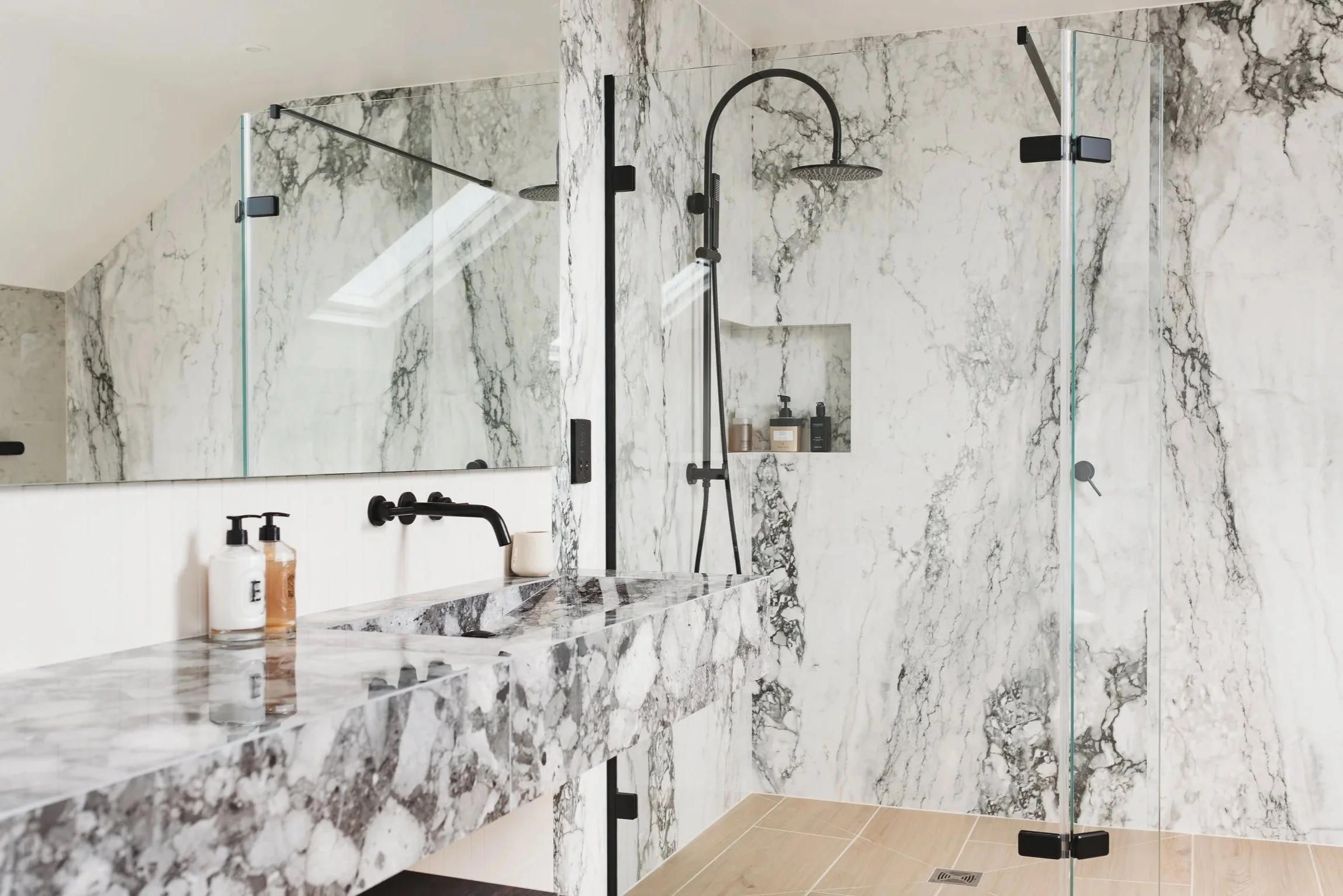 A modern luxury  bathroom with marble effect porcelain walls and a marble effect porcelain countertop, featuring a large mirror, a black shower, soap dispensers, and a glass-enclosed shower area with a rainfall shower head.