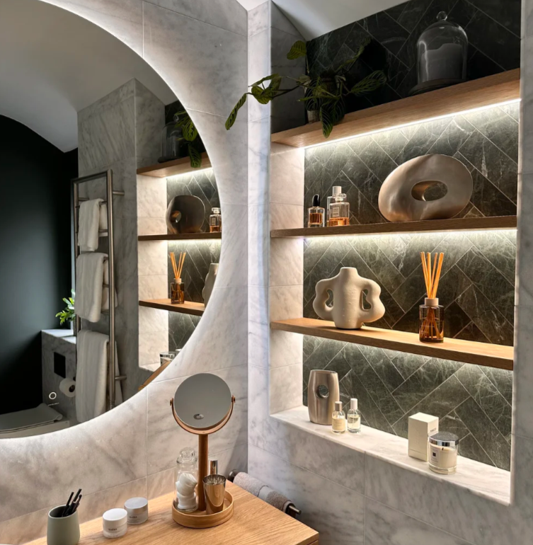 A bathroom vanity area with a large round mirror, wooden shelves displaying decorative vases, bottles, and reed diffusers, and a tidy wooden tabletop with skincare products and a mirror.