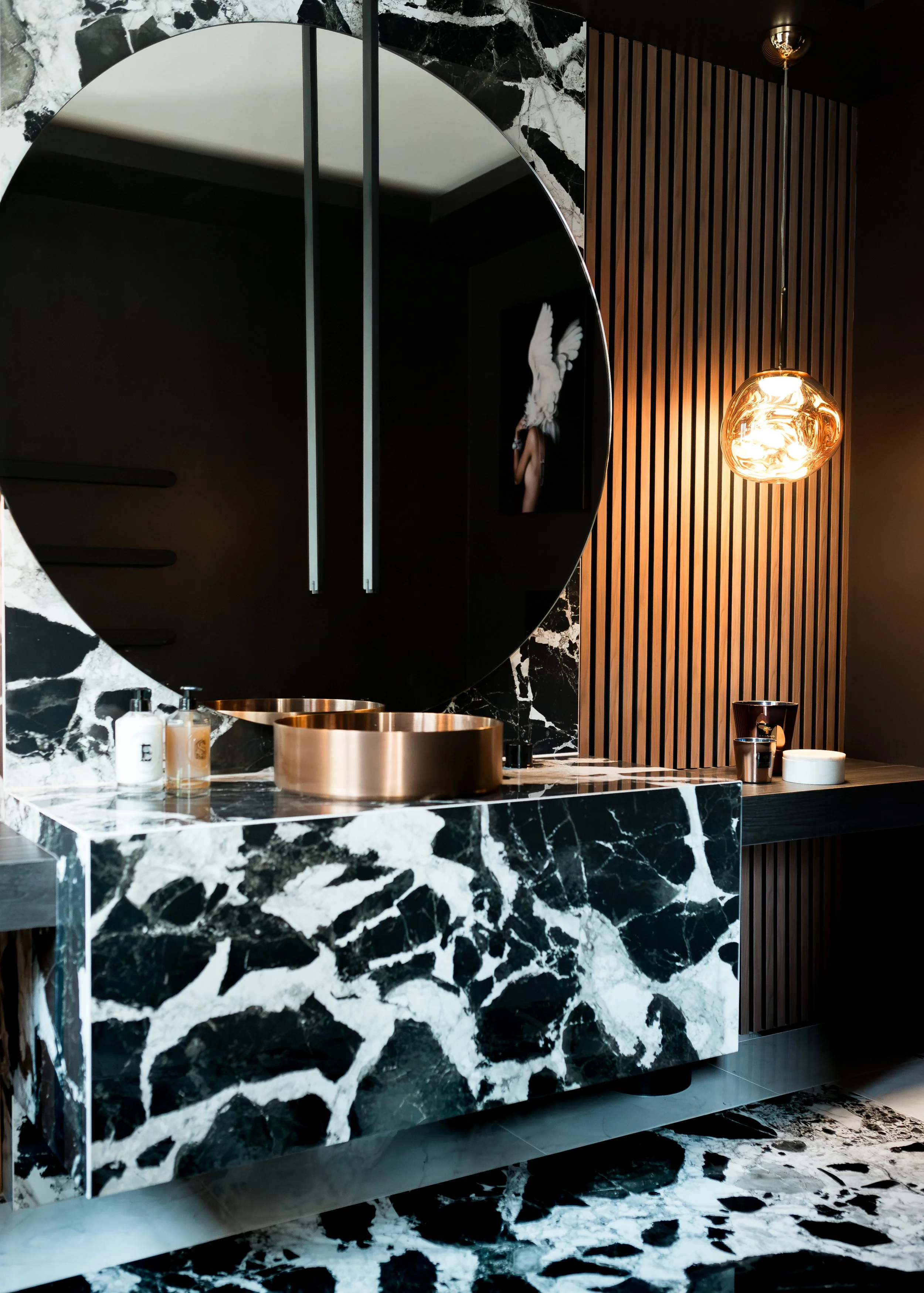 Luxury cloakroom bathroom. Cow print large format tile with luxury bespoke vanity with brass counter top basin. Extra large bespoke round mirror with luxury wood panelled walls to the side. Single glass pendant.