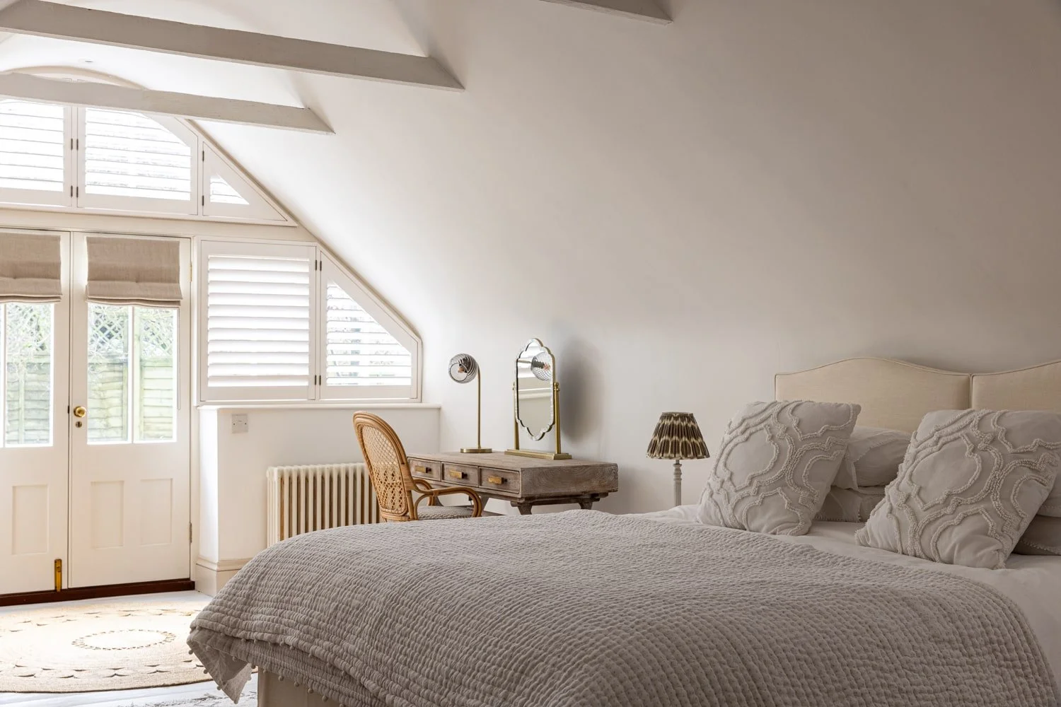 A cozy bedroom with a white bed, decorative pillows, a vintage dresser with a mirror, a small lamp, and a window with blinds letting in natural light.