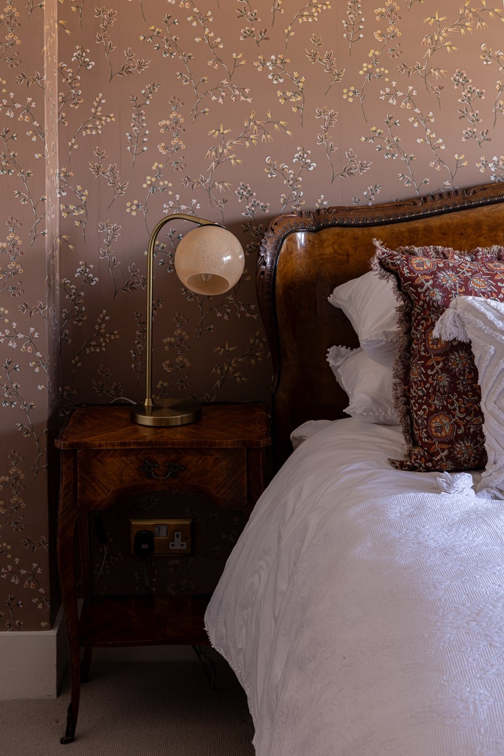 Close-up of a bedroom corner featuring a wooden nightstand with a beige modern lamp, a floral wallpapered wall, and part of a bed with white bedding and decorative pillows.