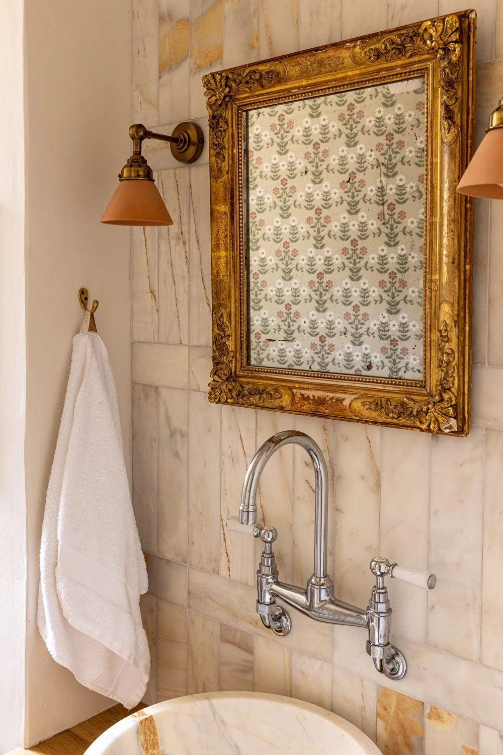 Bathroom with marble tiles, a gold ornate framed mirror, two wall-mounted sconces with tan shades, a towel hanging on a hook, and a polished chrome faucet above a marble sink.