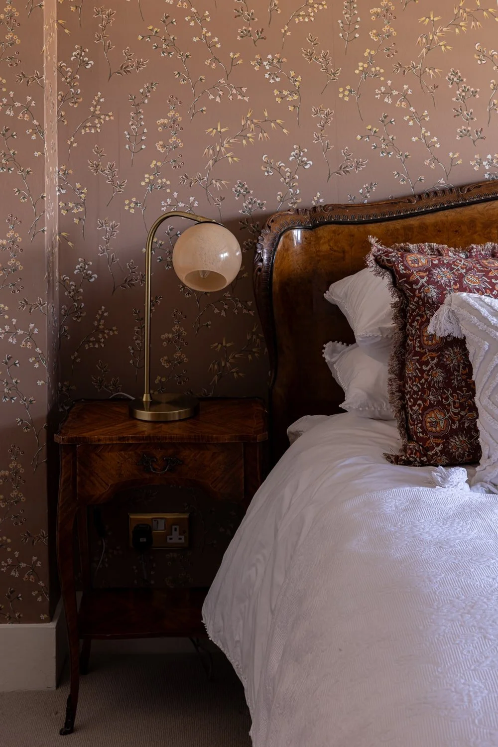 Close-up of a bedroom corner with a wooden headboard, patterned cushions, a white sheet, a wooden bedside table with a lamp, and floral wallpaper.