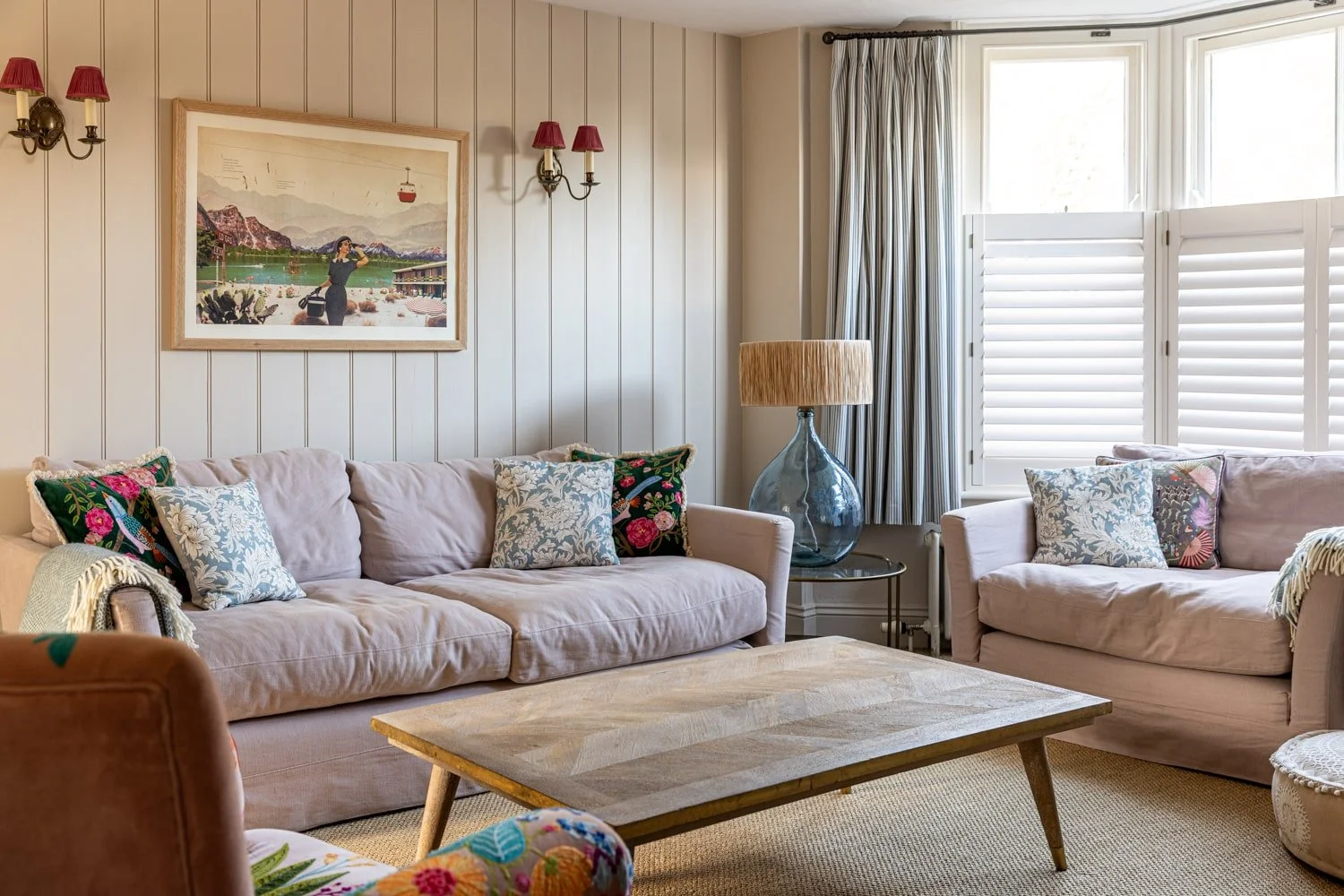 Living room with beige sofas, decorative pillows, a wooden coffee table, large window with shutters, a glass table with a large blue lamp, striped curtains, framed artwork, and wall-mounted sconces with pink lampshades.