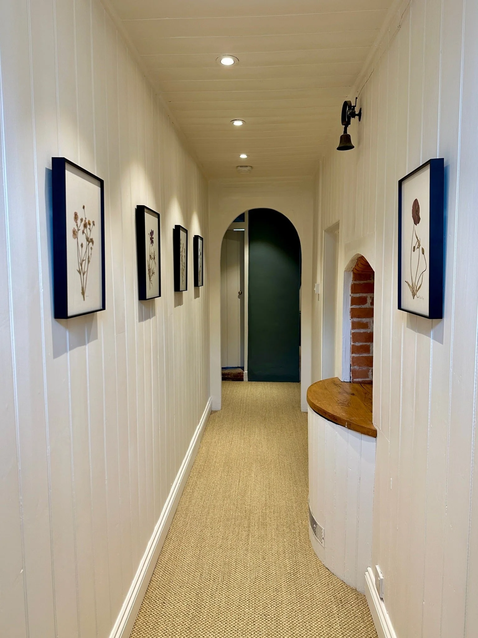 A hallway with cream-colored wood-paneled walls, carpeted floor, framed botanical art on the walls, and a black wall-mounted light fixture.