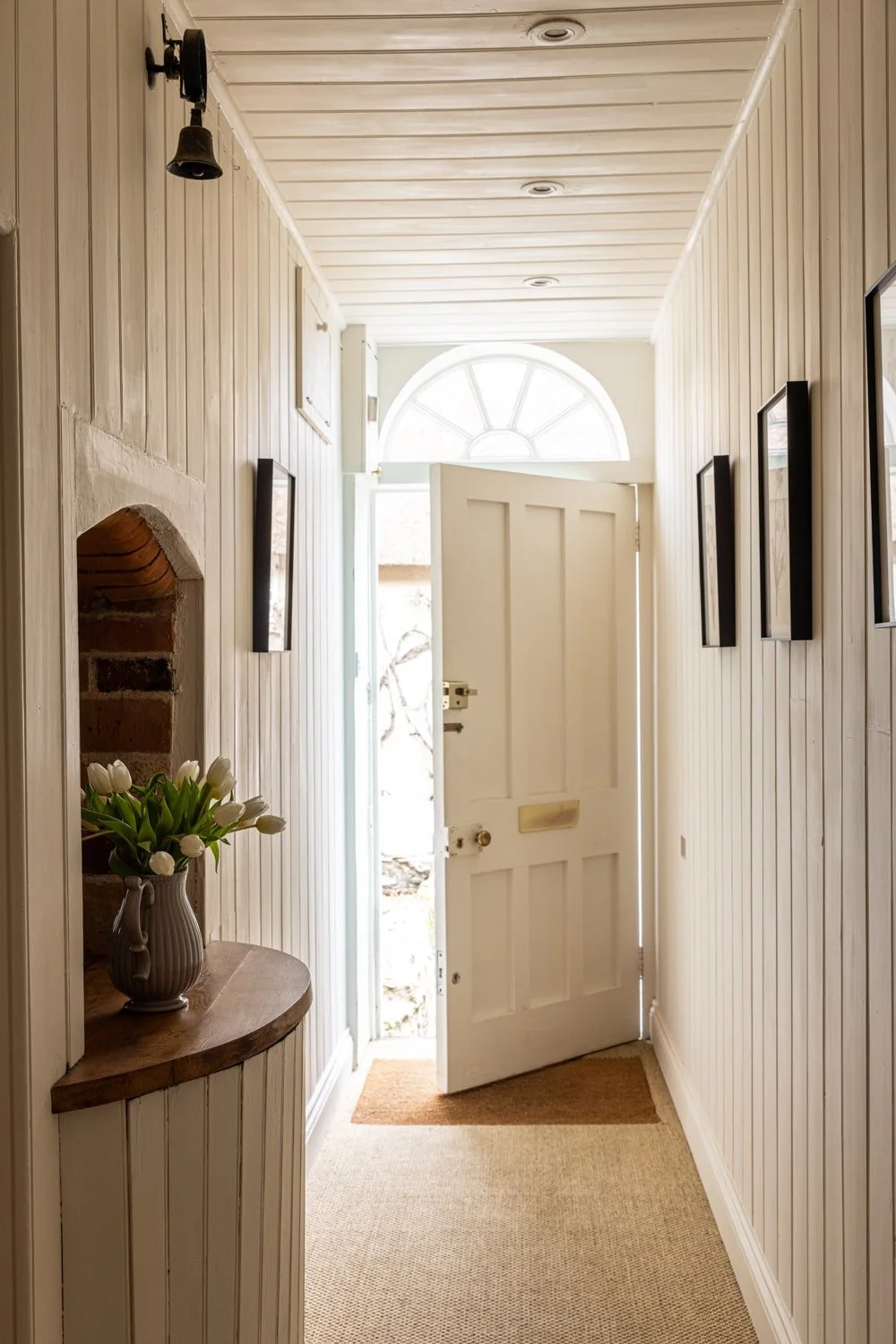 A bright narrow entryway with beige paneled walls, a white wooden door slightly open, a semi-circular window above, framed pictures on the walls, a small shelf with a vase of white tulips, and a doormat on a beige carpet.