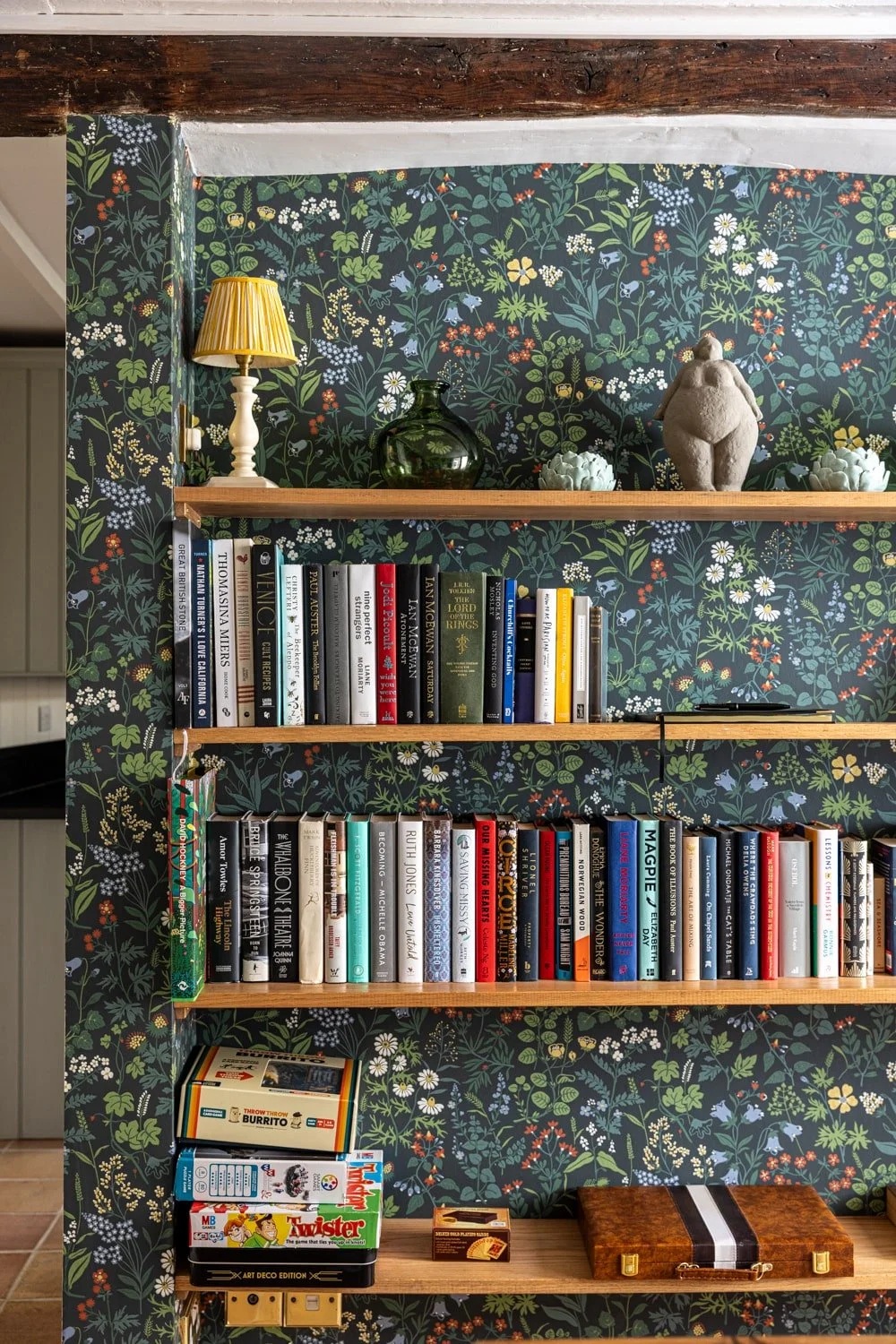 Bookshelf with three wooden shelves against a floral wallpapered wall. The top shelf has a yellow lampshade and decorative objects. The middle shelf has a collection of books. The bottom shelf has more books, board games, and a wooden briefcase.