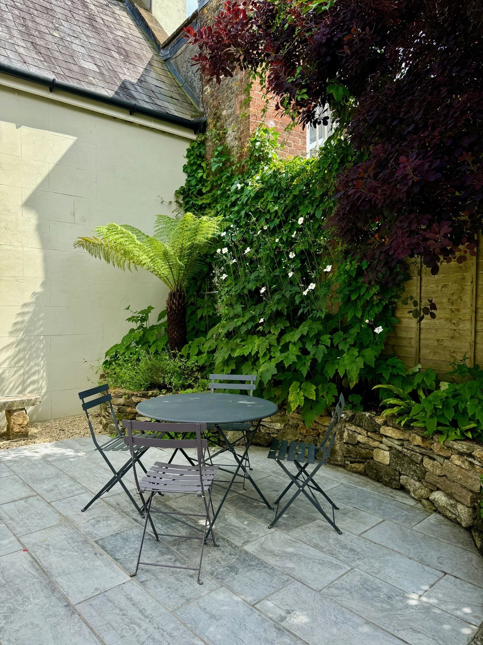 Small outdoor patio with four metal chairs around a round table, surrounded by greenery including a large fern, flowering vines, and a purple-leafed tree, with stone pavement and a stone border.