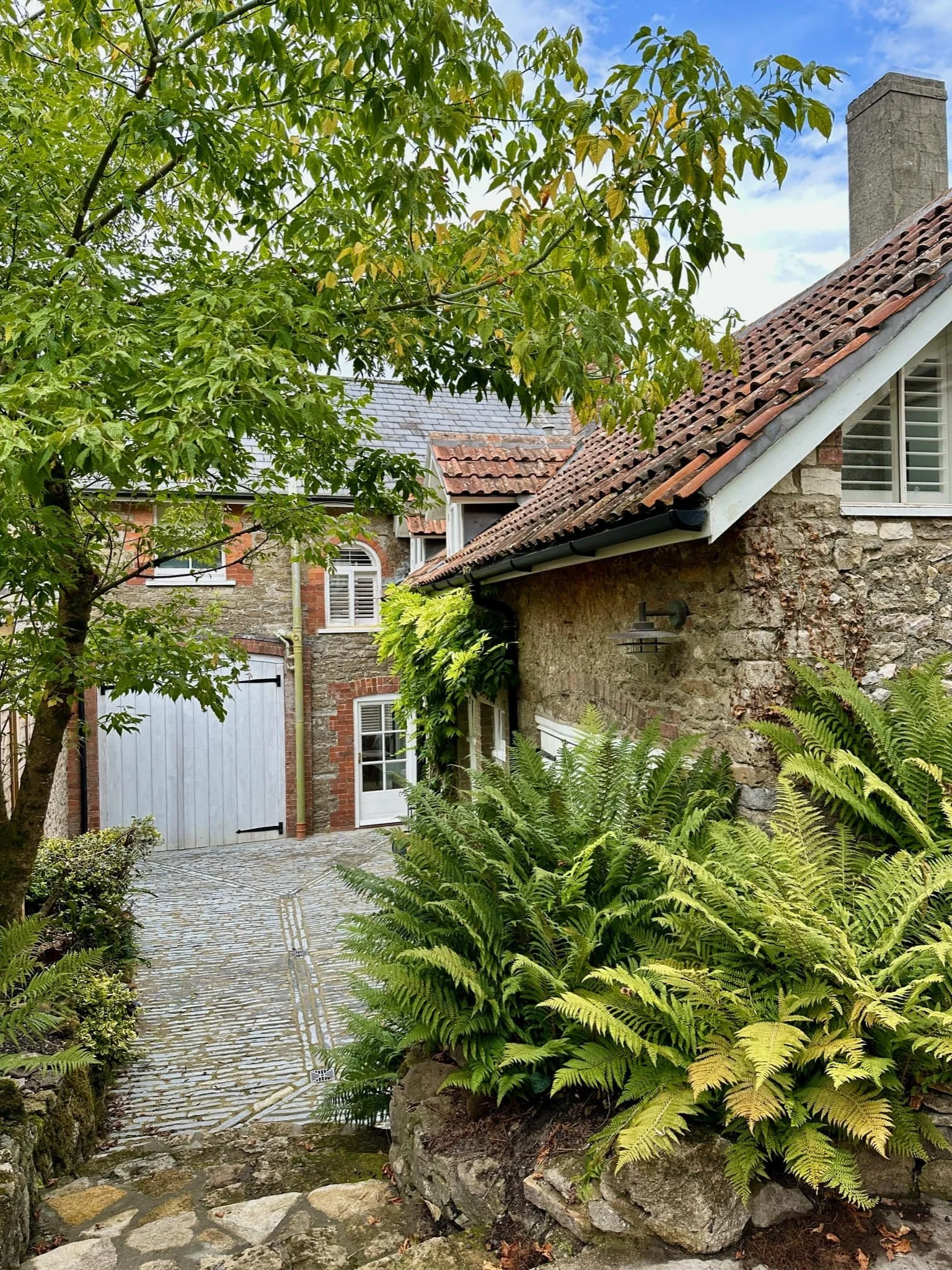 A stone cottage with a tile roof, surrounded by greenery and a cobblestone path.
