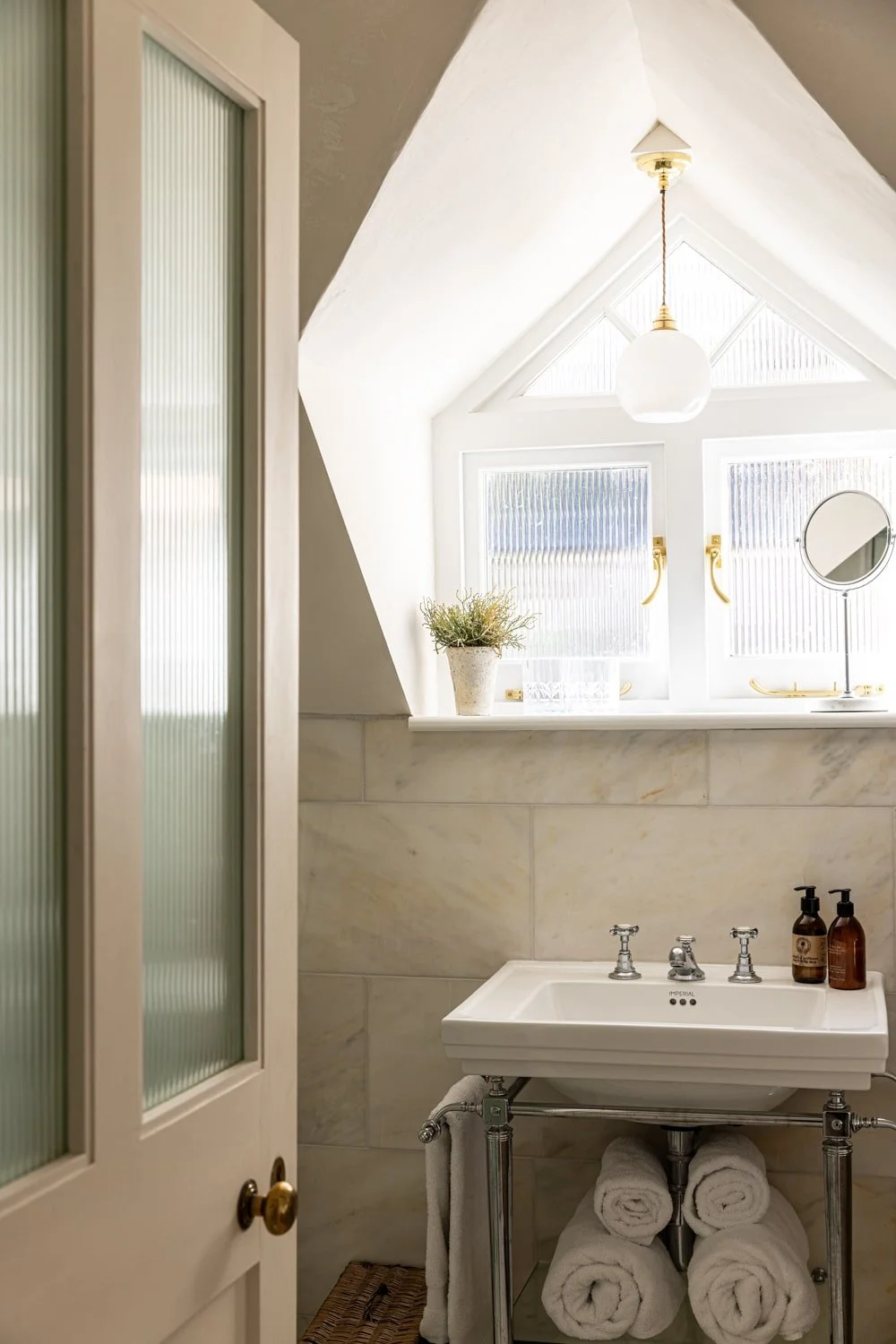 A small bathroom with a white ceramic sink, rolled white towels underneath, a window with frosted glass, a potted plant on the windowsill, and brushed gold fixtures. Light filters through a triangular window with a hanging pendant light.