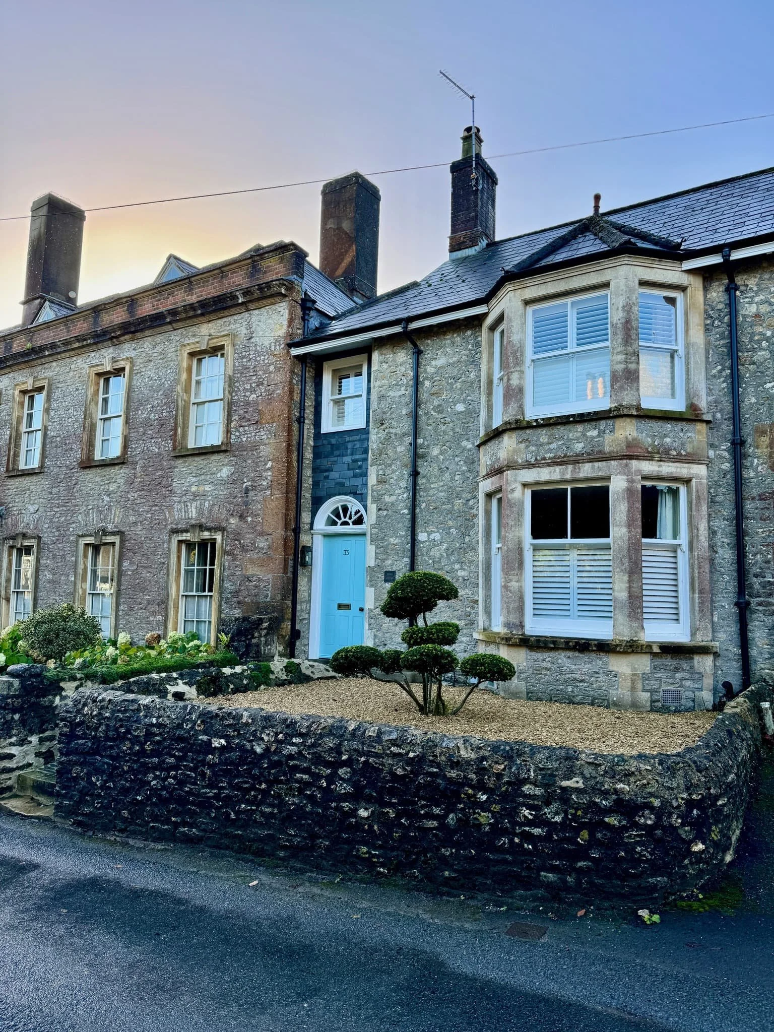 A stone building with a blue front door and large bay windows with white shutters. A small, manicured tree is in front of the house. The building has multiple brick chimneys and a sloped roof, and the sky is clear.