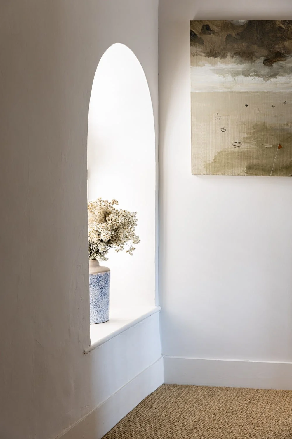 Decorative white vase with dried flowers on a white windowsill with an arched window, accompanied by a painting with cloudy sky and boats on the wall.