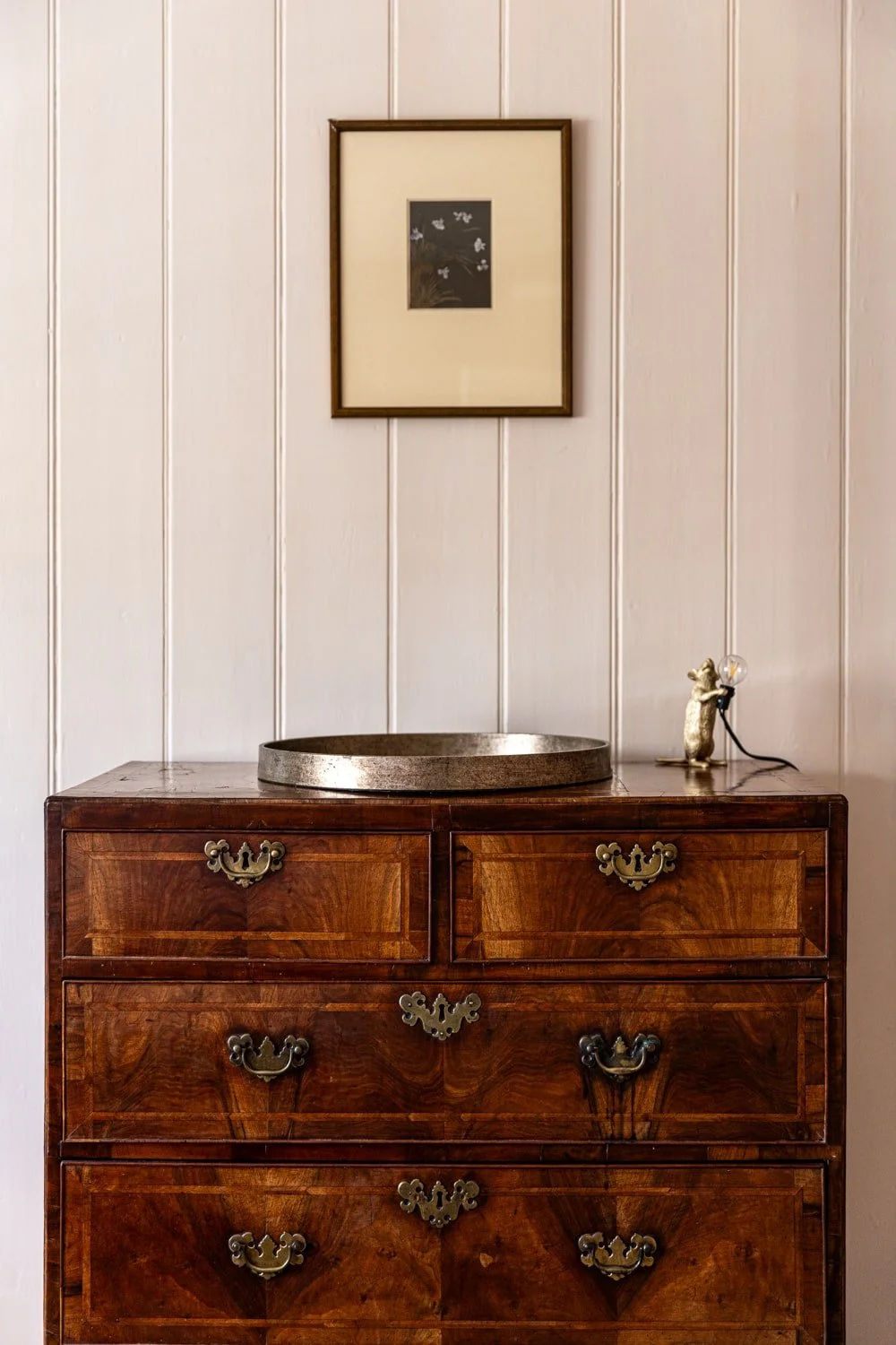 A vintage wooden dresser with brass handles, a round metallic tray on top, a small dog figurine holding a light bulb, and a framed artwork of white butterflies on a dark background hanging above on white paneled wall.