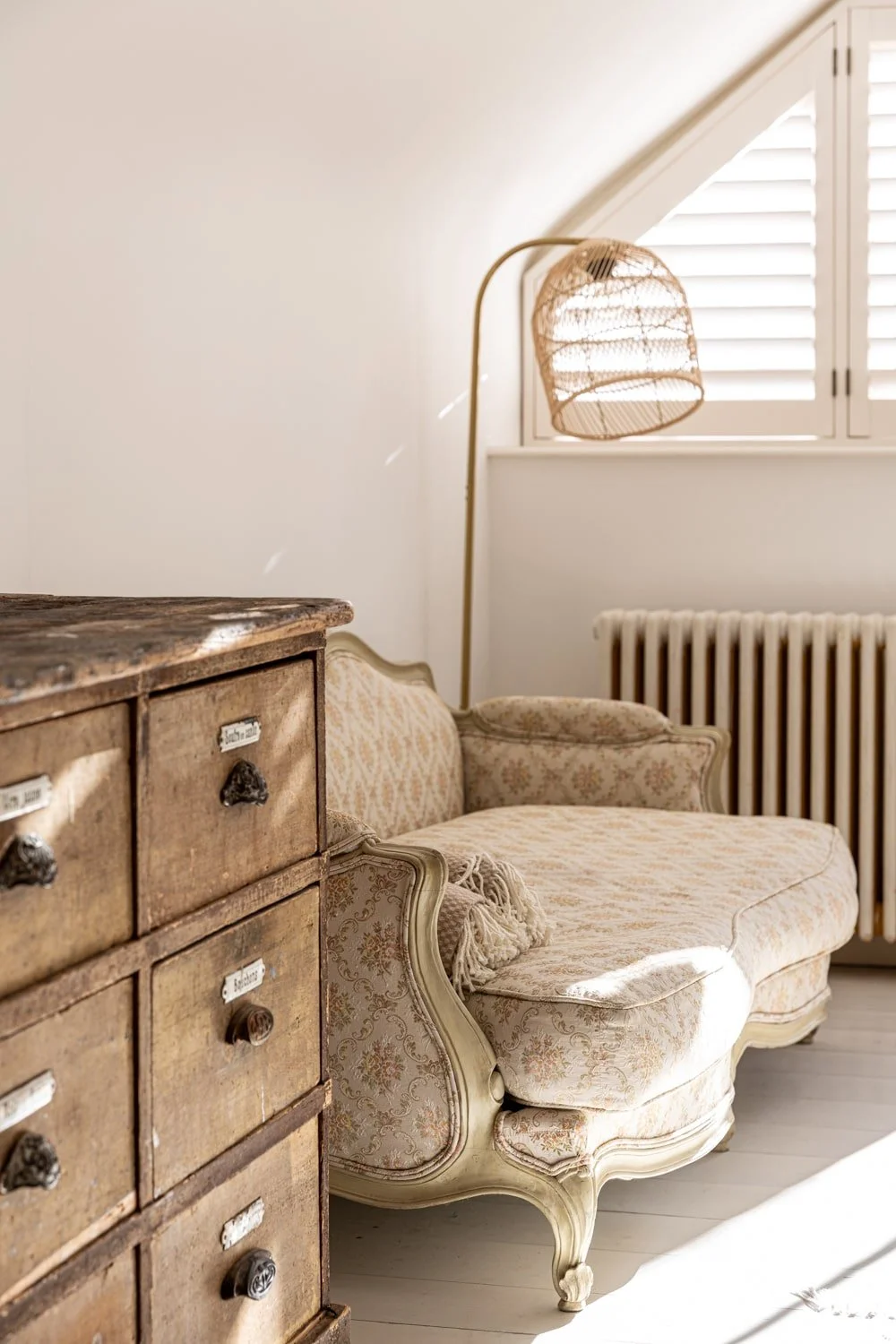 A vintage cream-colored sofa with floral upholstery next to an old wooden chest of drawers with labels on the drawers. Behind the sofa is a window with white plantation shutters and a brass floor lamp with a woven rattan shade.