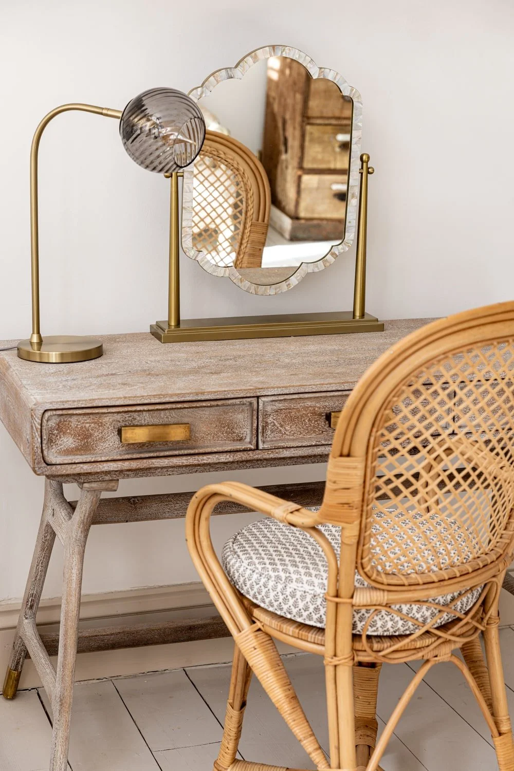 A vintage wooden desk with a distressed finish, a round brass-handled drawer, and a brass table lamp. A decorative mirror with shell-like edges and ruffled glass is placed on the desk. There is a rattan chair with a woven backrest and a patterned cus