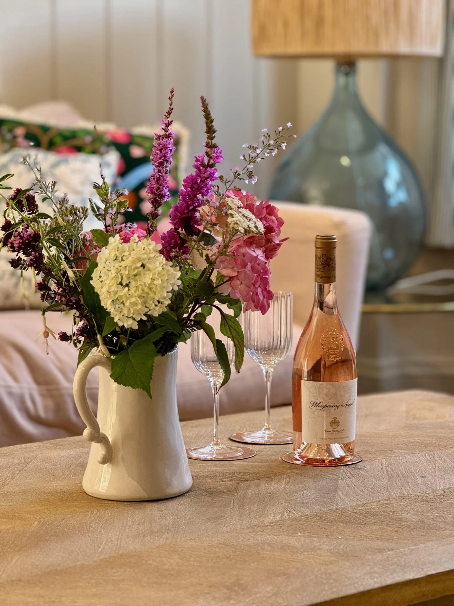A table with a bouquet of pink, white, and purple flowers in a white pitcher, two empty champagne flutes, and a bottle of rosé wine labeled "Whispering Angel" in a cozy, well-lit room.