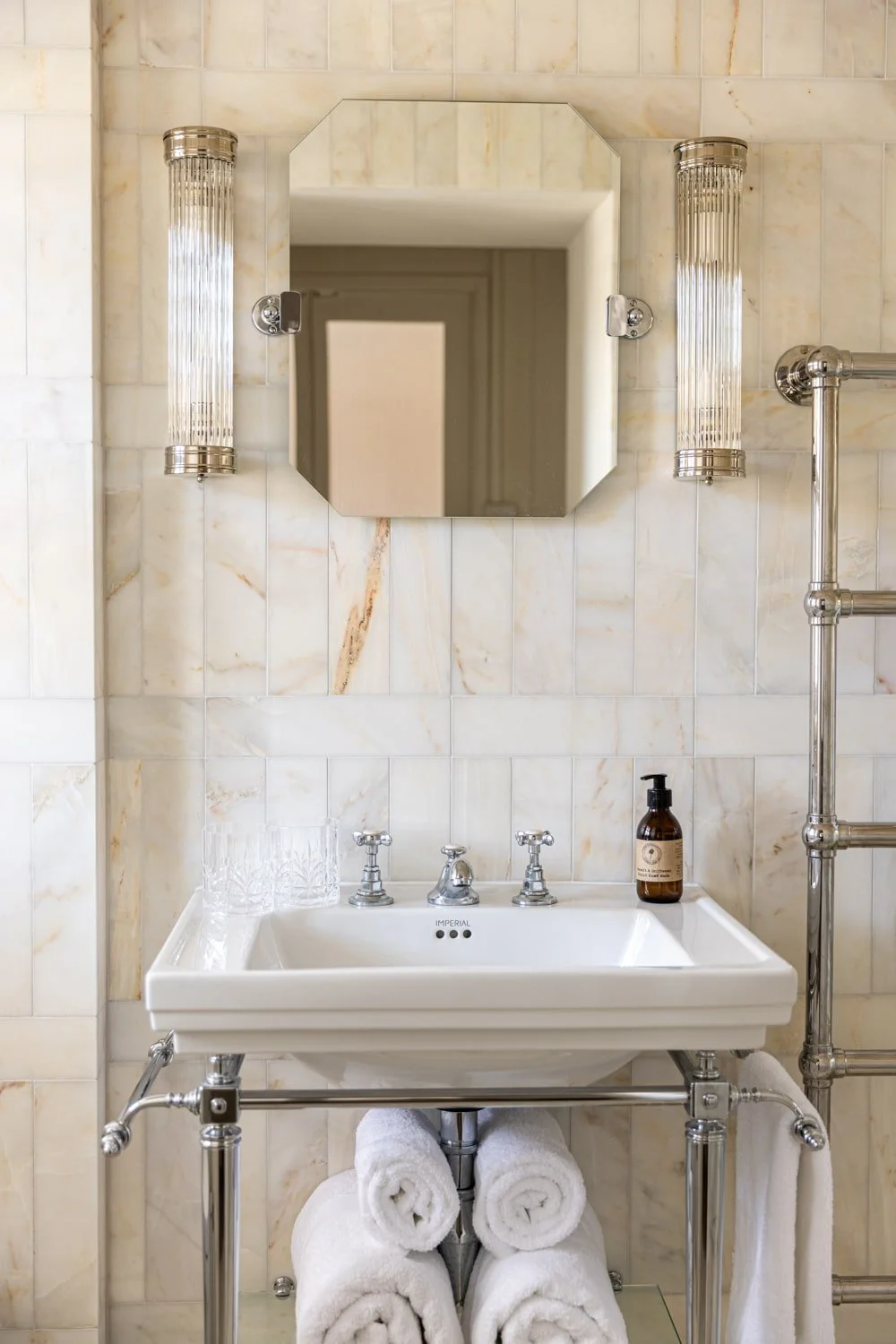 Bathroom sink with a mirror, two wall sconces, and three rolled white towels stored underneath, with a bottle of soap on the sink, and a marble tiled wall.