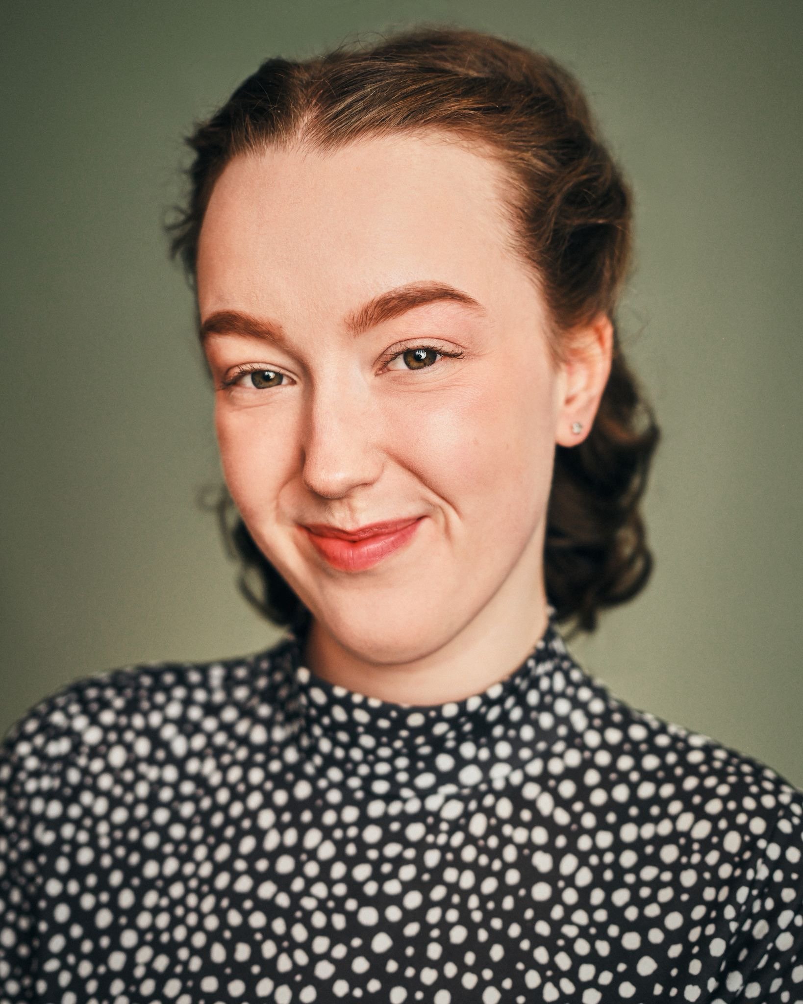 A young woman with curly hair, smiling, wearing a black shirt and a blue ribbon in her hair, standing against a plain white background.