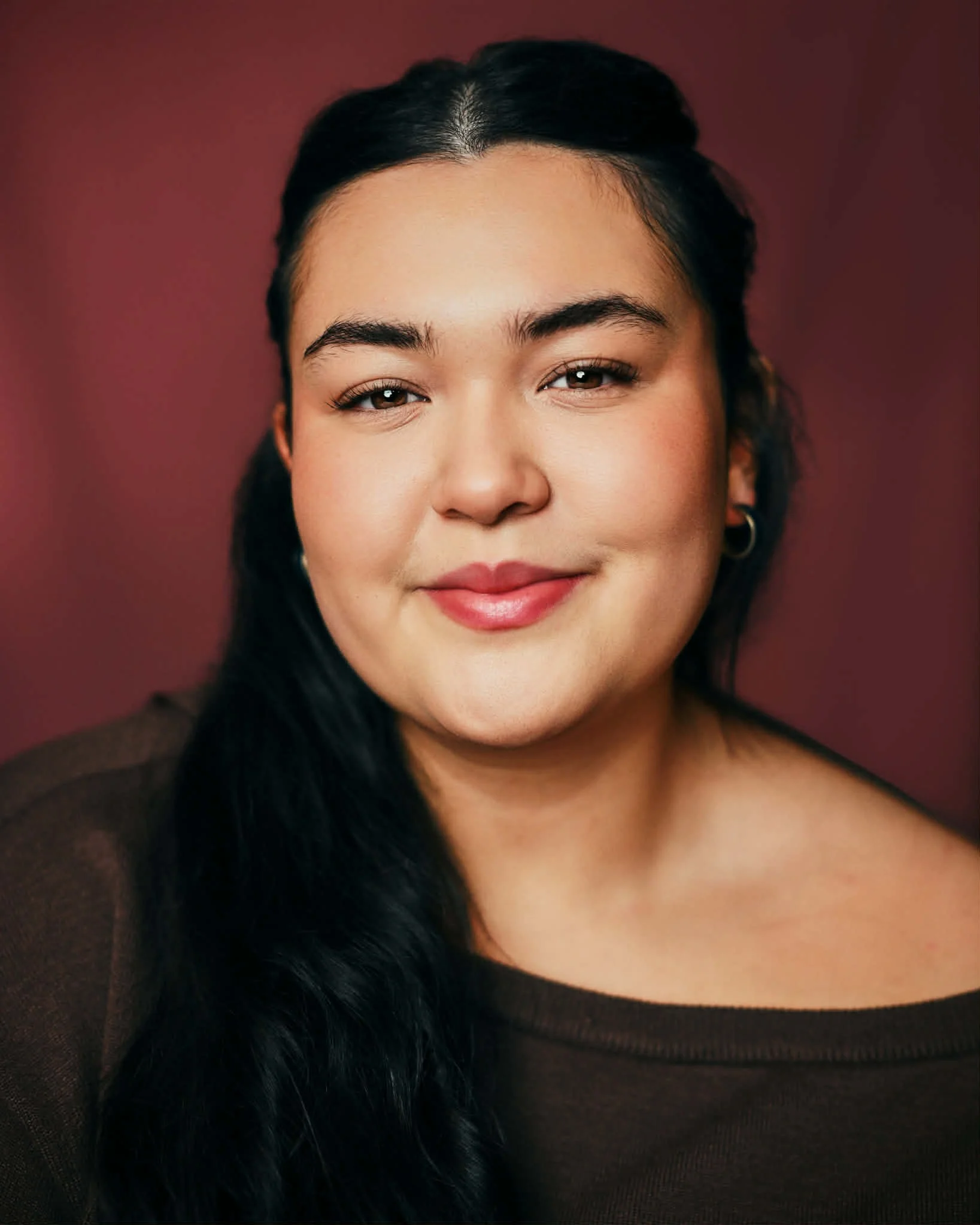 Young woman with long black curly hair smiling, wearing a red long-sleeve top against a plain light-colored wall.