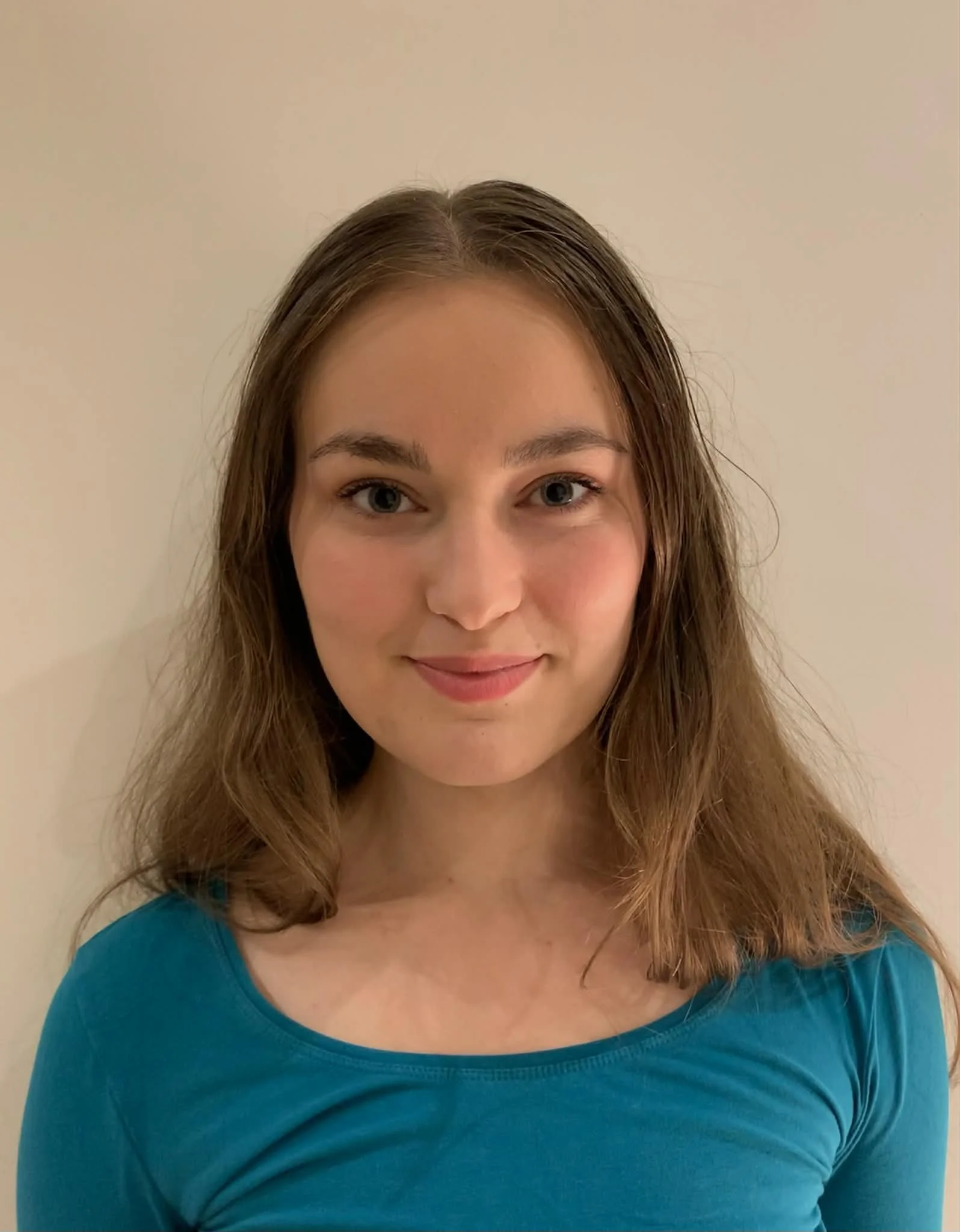 A young woman with long brown hair tied back with a blue scrunchie, smiling, wearing a white knit sweater and a pearl necklace, standing against a plain white wall.