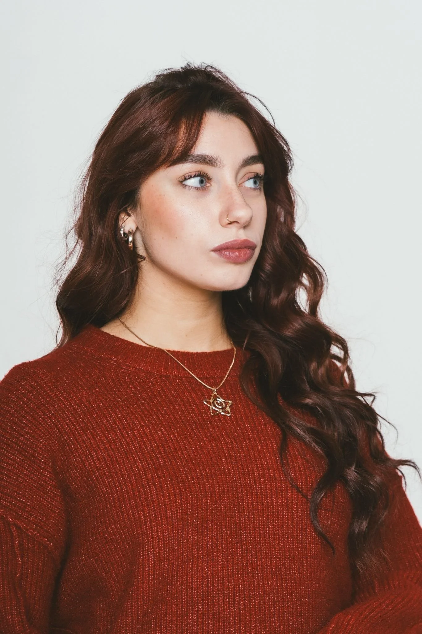 A young woman with long, wavy red hair and a fair complexion, wearing a light gray long-sleeve shirt and gold jewelry, standing in front of a plain white wall.