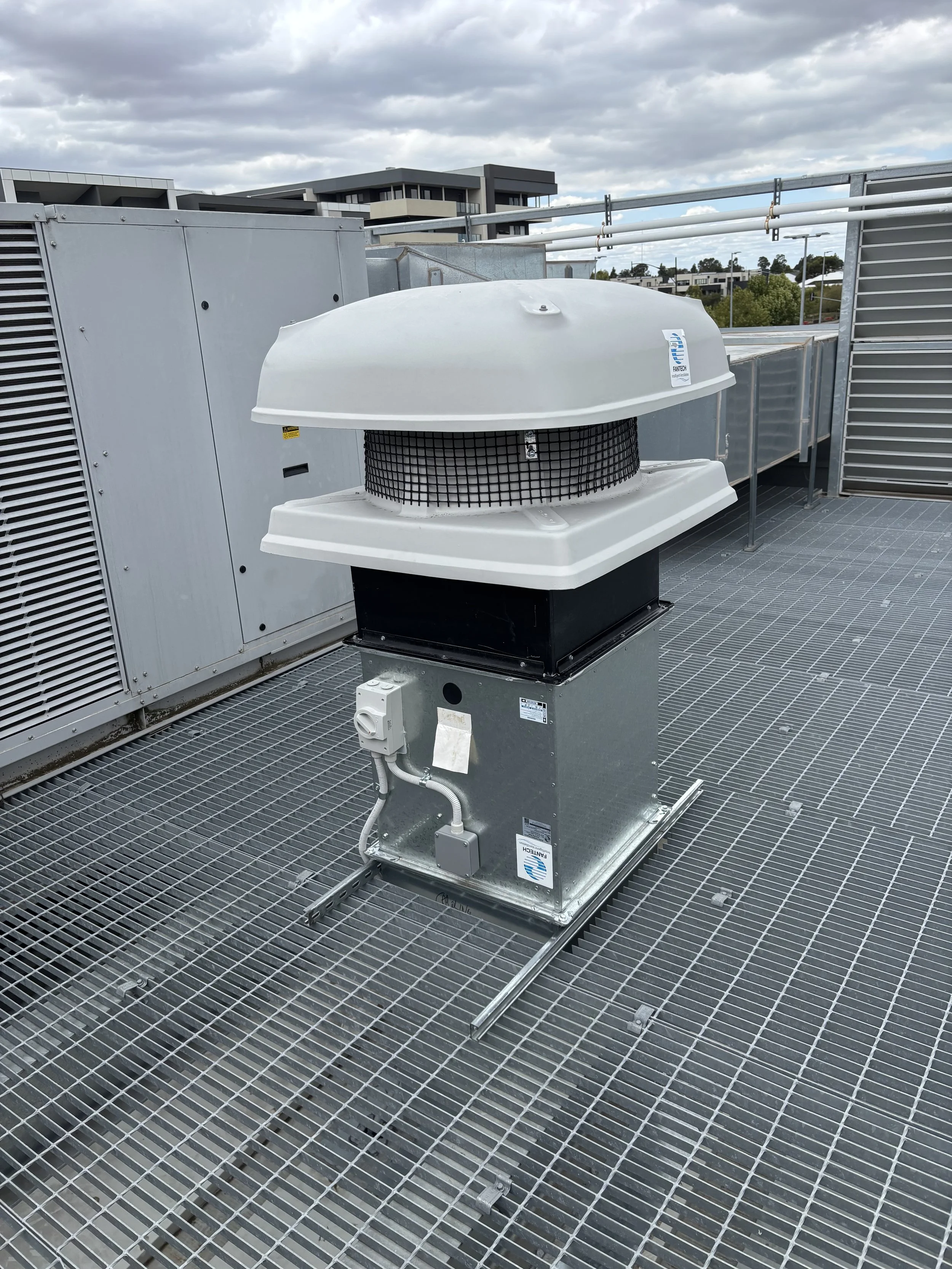 Roof-mounted ventilation unit on metal grating platform with urban buildings in the background.