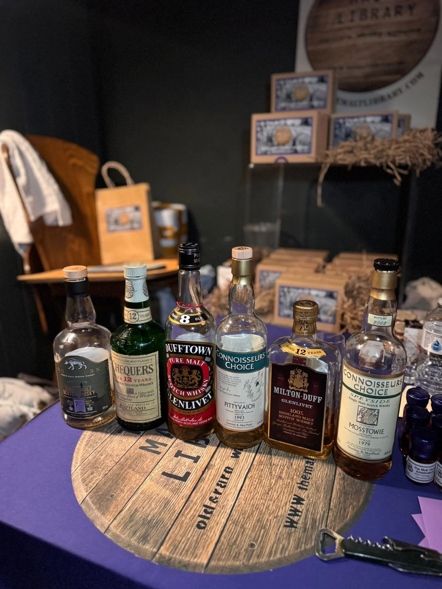 Six bottles of whisky displayed on a wooden table at a tasting event, with various labels including Connoisseurs Choice, Dufftown, and Milton-Duff, in a dimly lit room with promotional materials and boxes in the background.