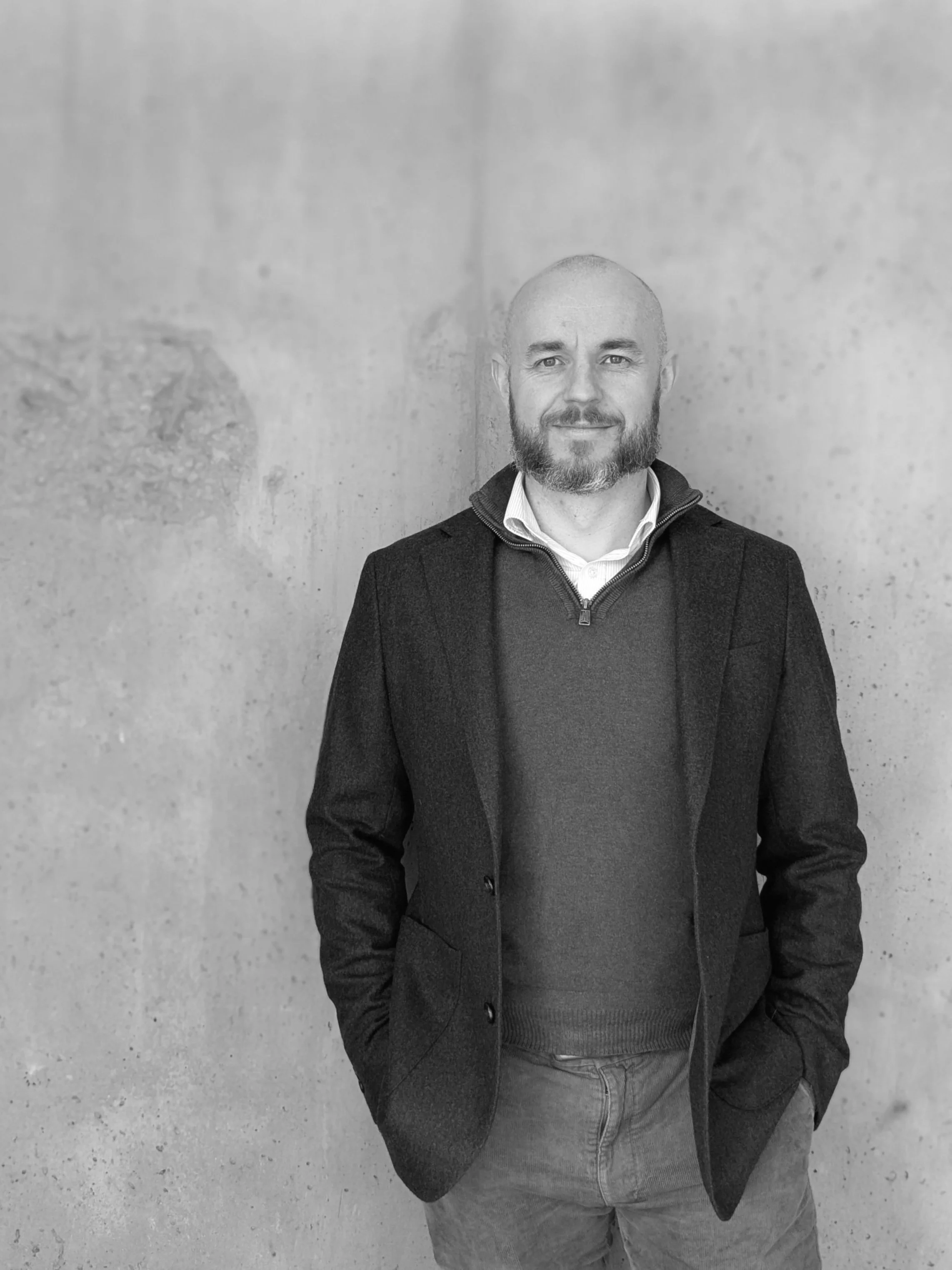 Black and white photo of a man in a blazer and sweater, standing against a concrete wall.