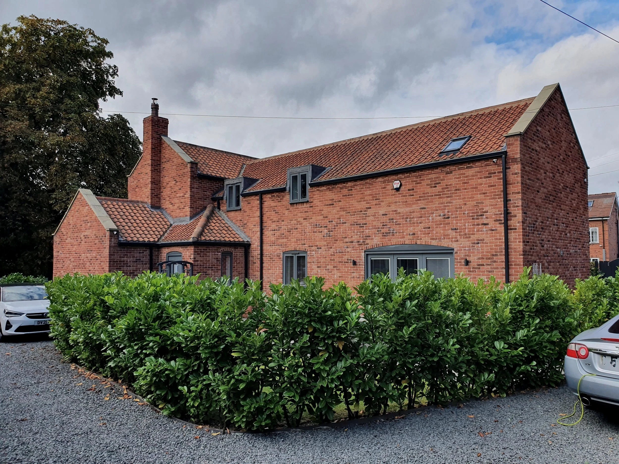Brick house with pitched roof and hedges, cars parked on gravel driveway