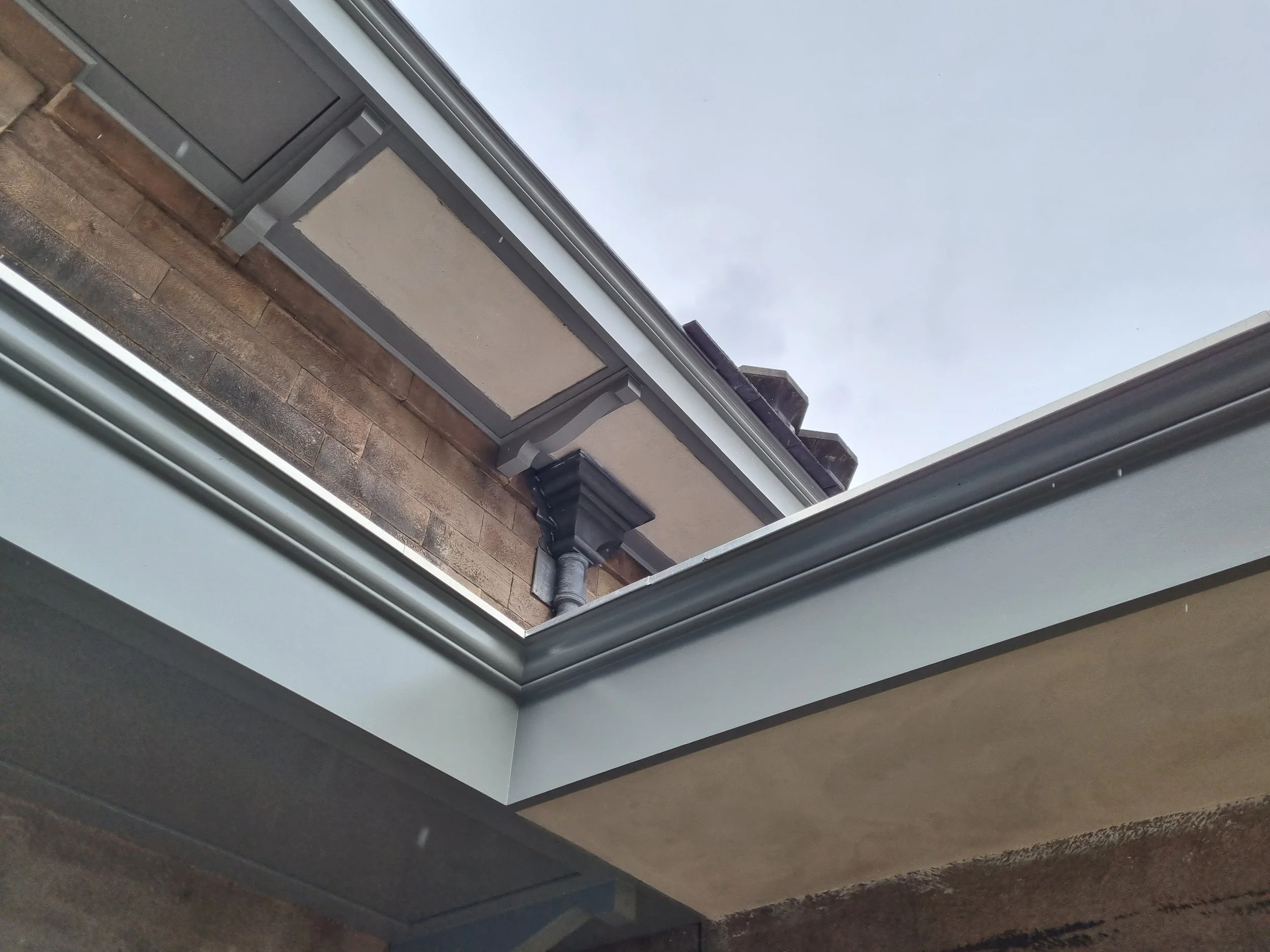 View looking up at a building's gutter system and roof, featuring a drainpipe against a brick wall and a cloudy sky background.