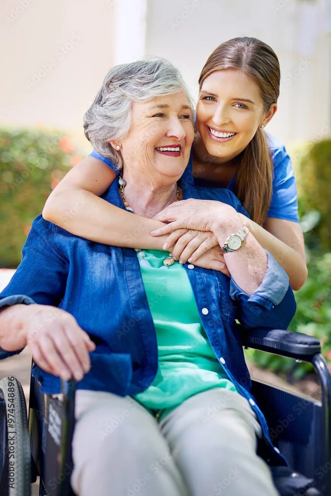 A smiling elderly woman in a wheelchair is embraced by a young female caregiver in an outdoor setting.