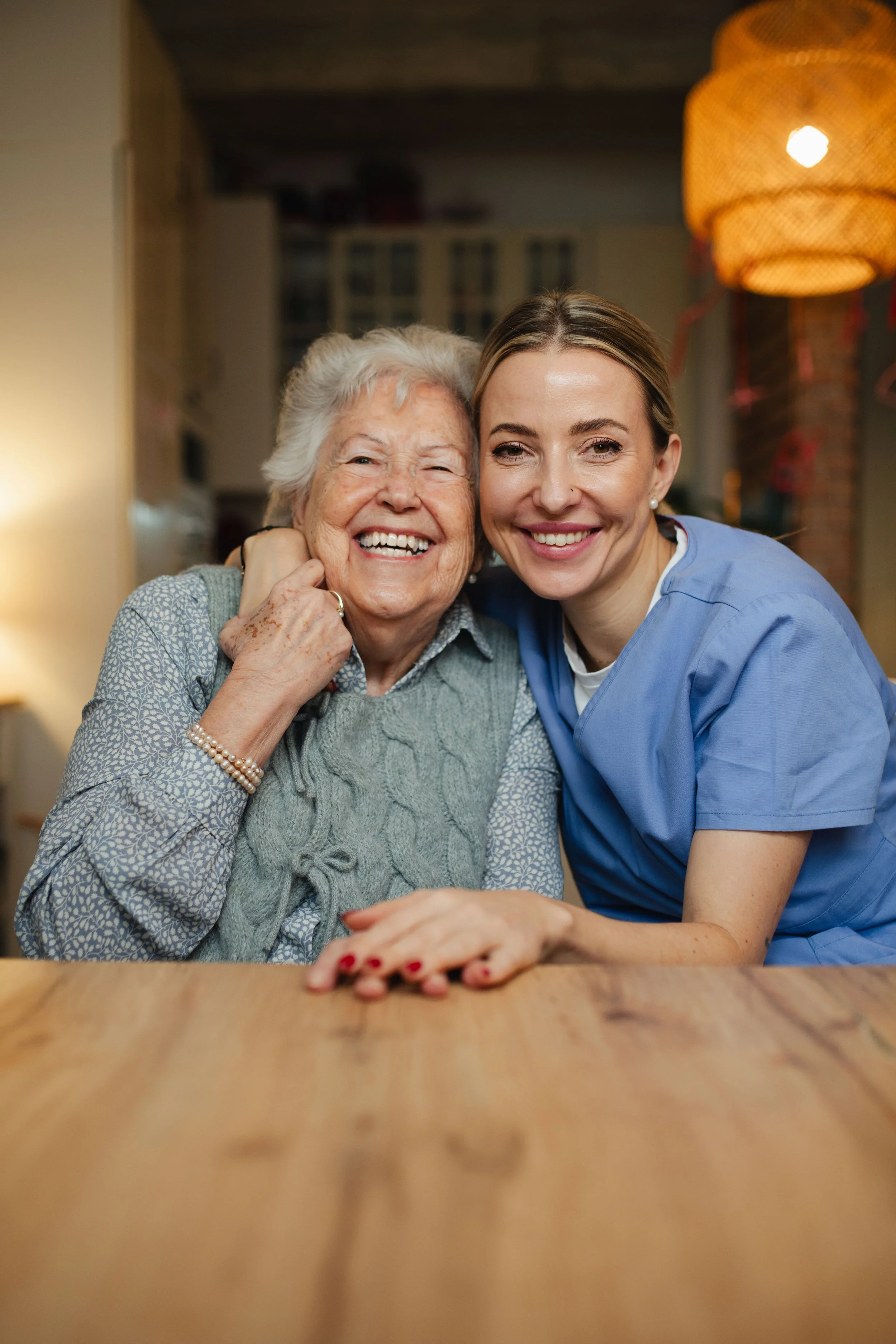 A smiling elderly woman and a woman in blue scrubs sitting together at a wooden table, embracing each other warmly in a cozy room.