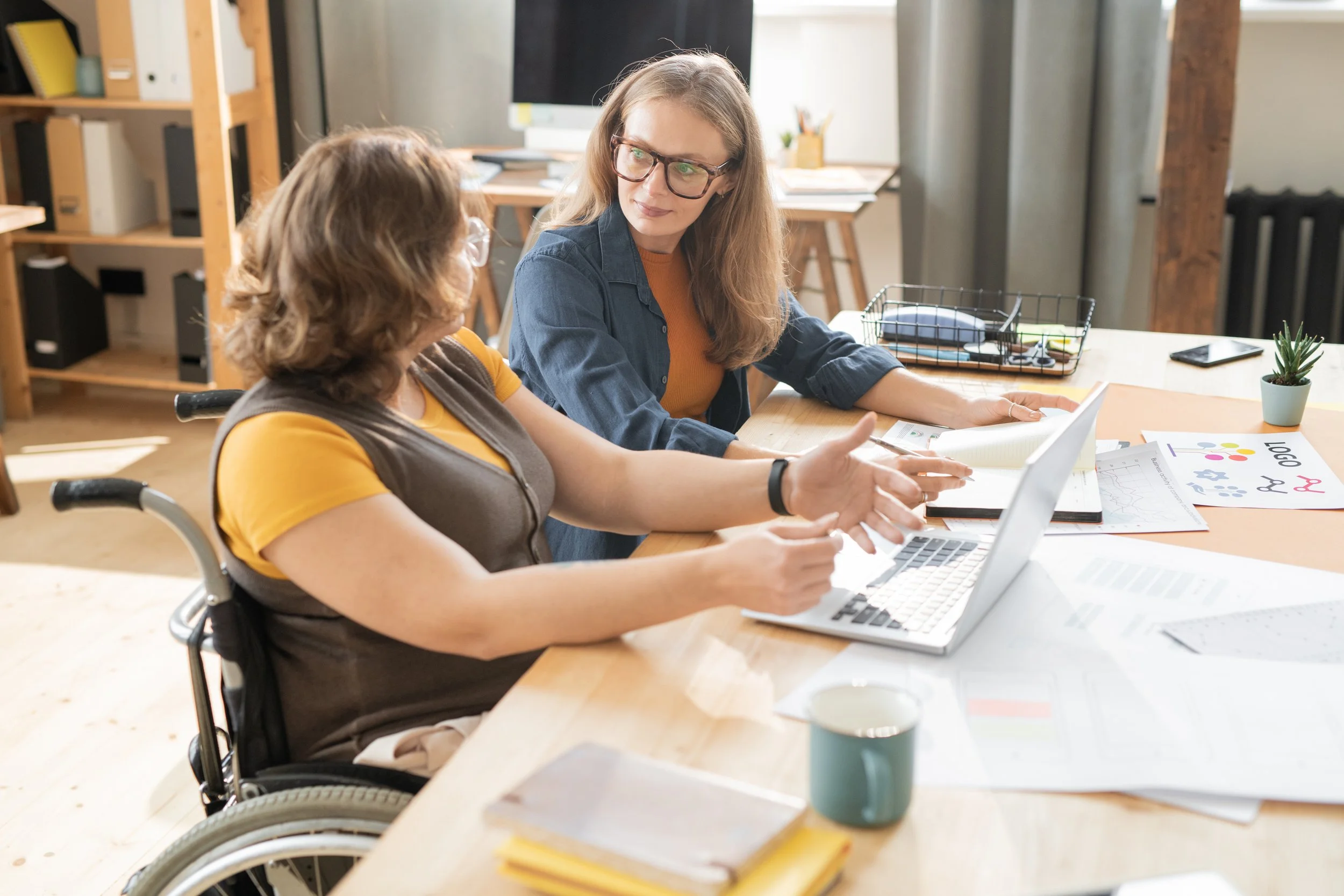 Two women in a meeting at a desk with a laptop, one in a wheelchair, discussing charts and documents.