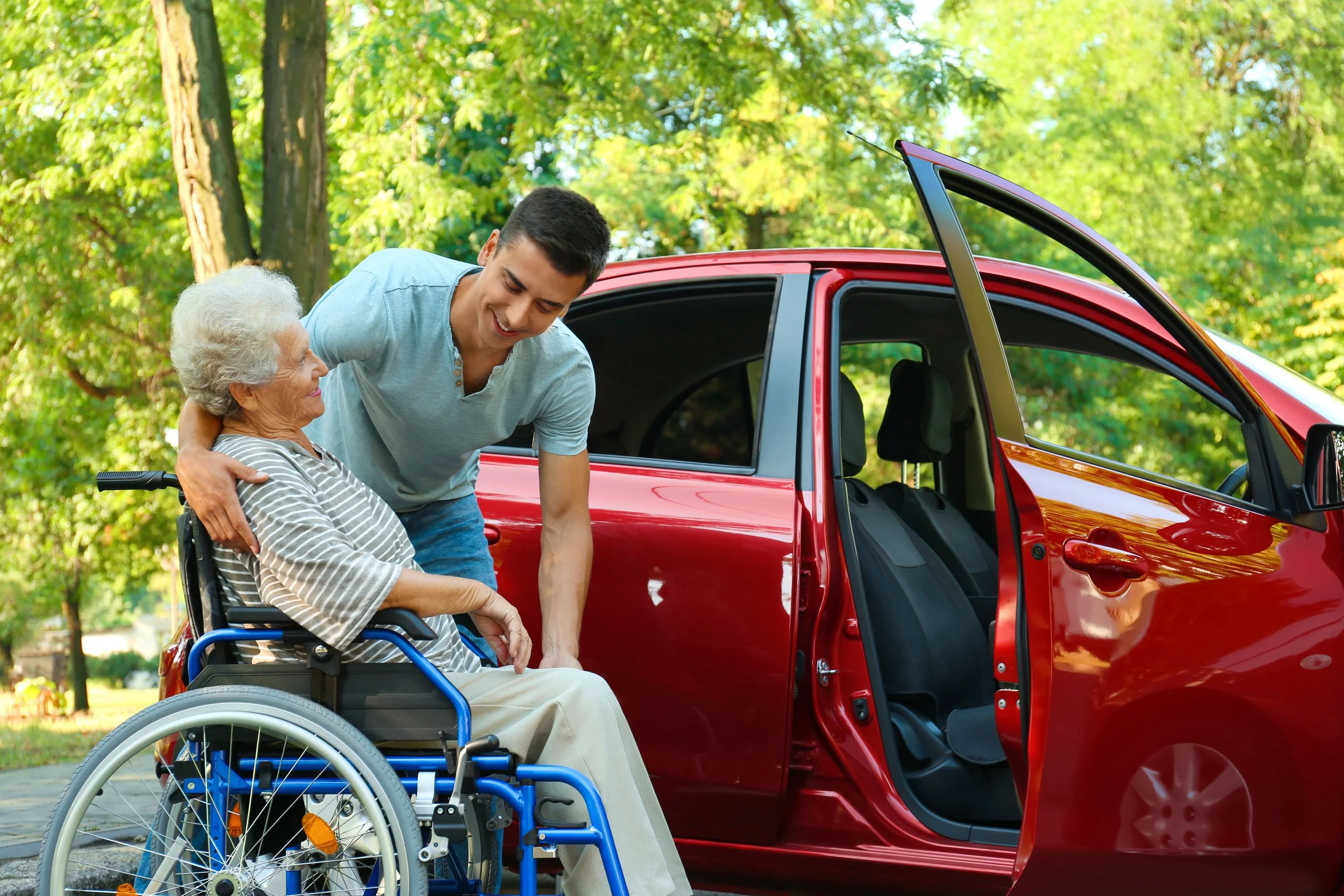 Young man assisting an elderly woman in a wheelchair near a red car with an open door in a park setting.